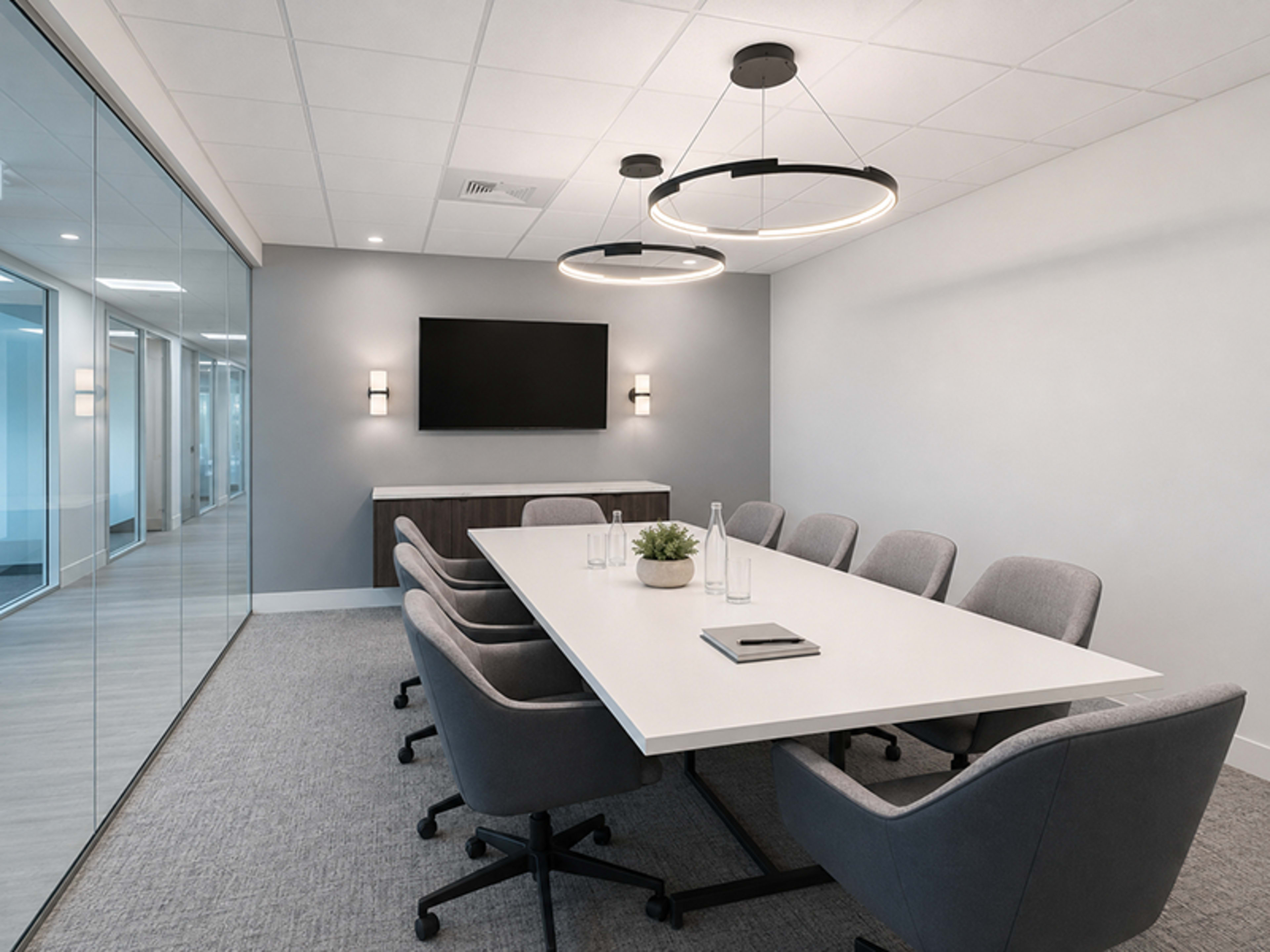 The image shows a modern conference room featuring a long table surrounded by ergonomic chairs, a wall-mounted TV, and sliding glass doors.