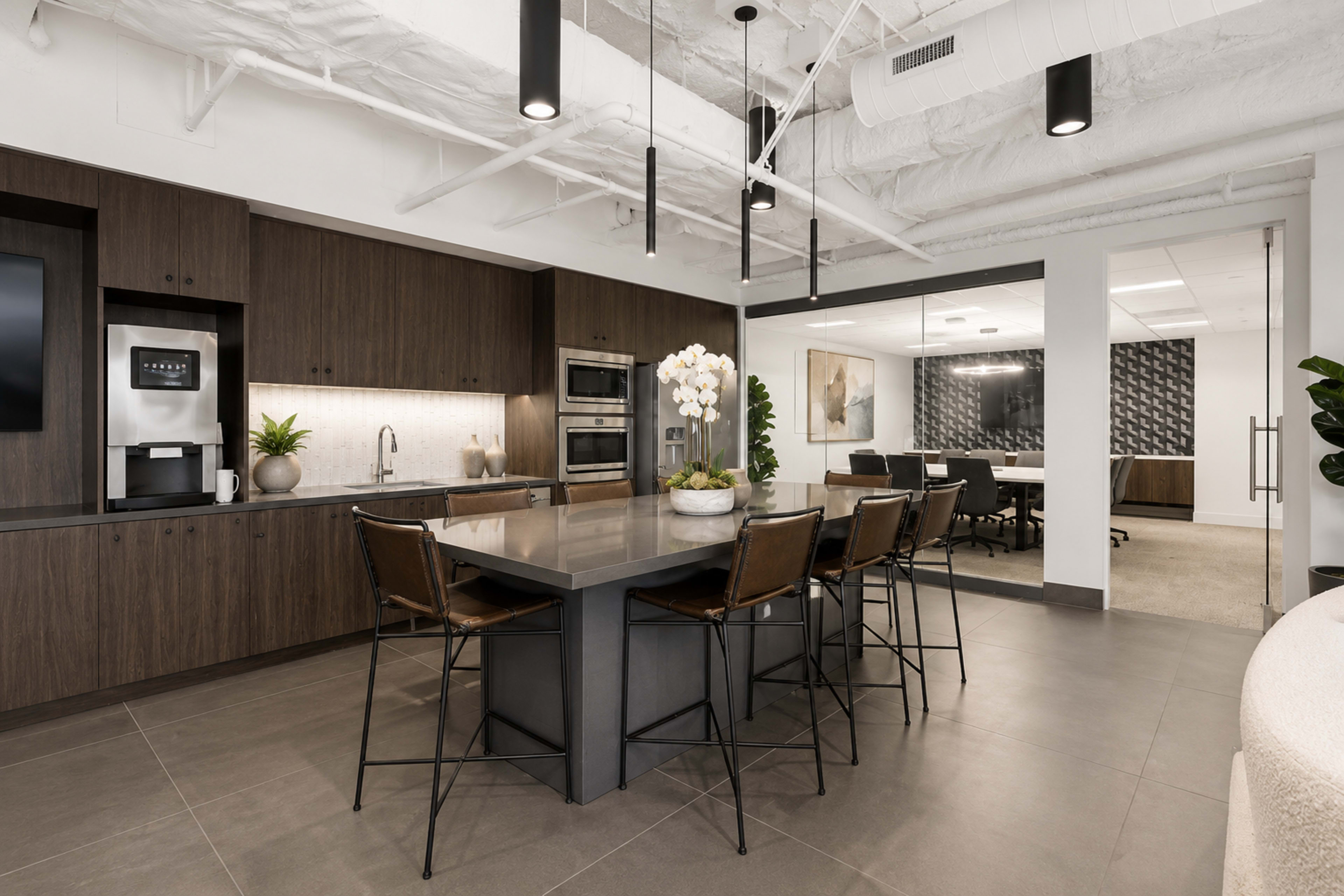 The image shows a modern kitchenette with dark wood cabinetry, a large island with seating, and a dining area leading into a conference room.