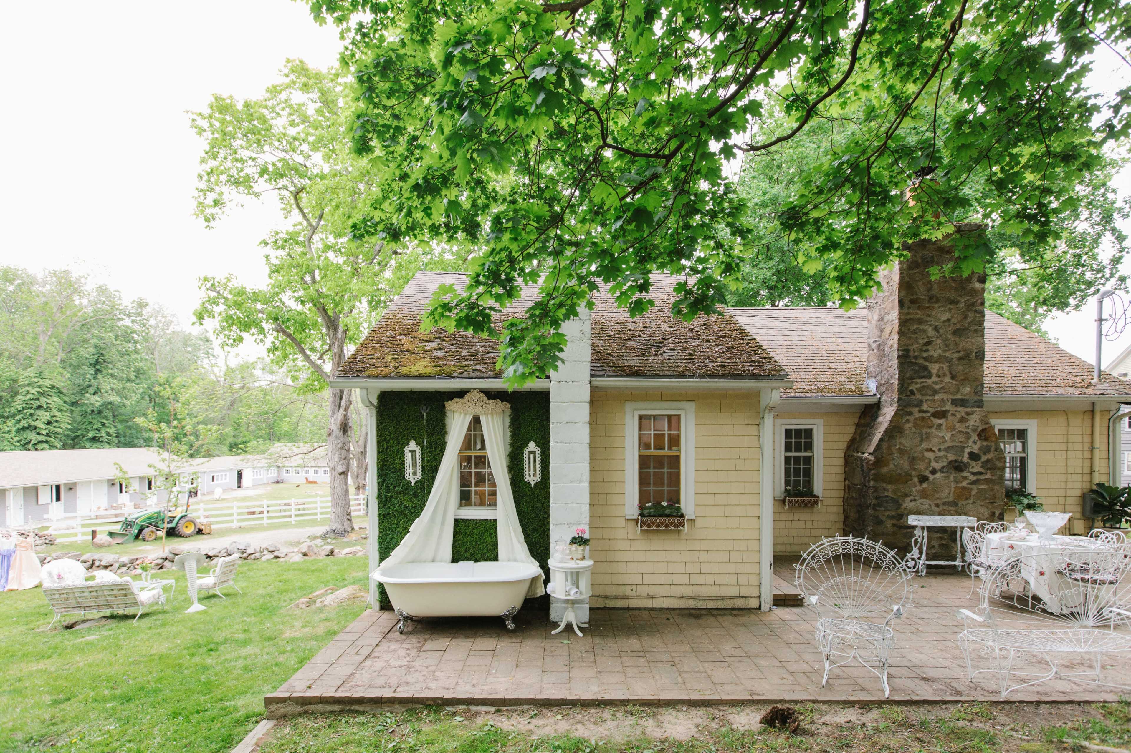 A quaint cottage is set amid green trees, featuring a vintage bathtub on the patio and a stone chimney alongside a grassy yard.