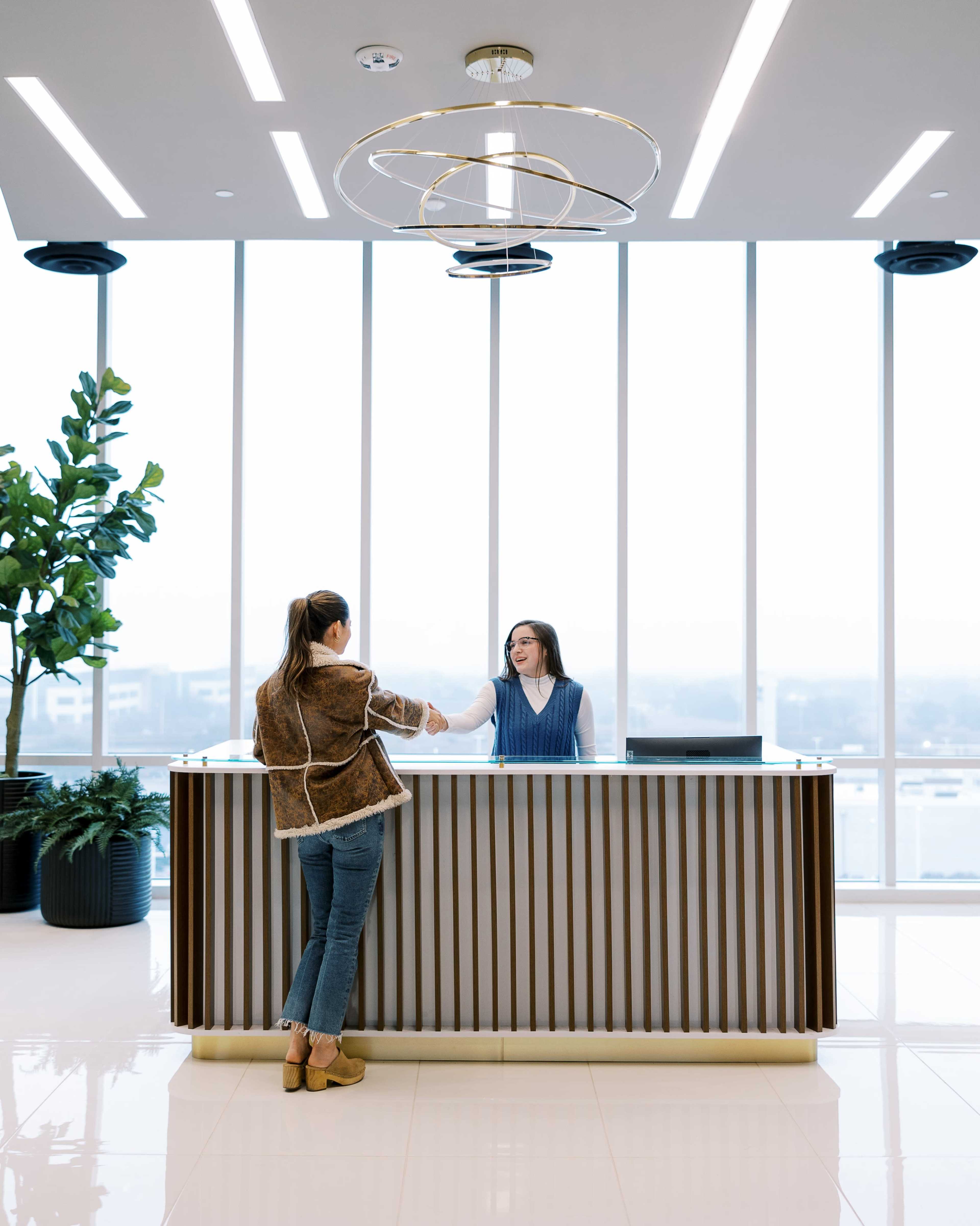 A woman in a brown jacket shakes hands with a receptionist behind a modern front desk in a brightly lit lobby with large windows.