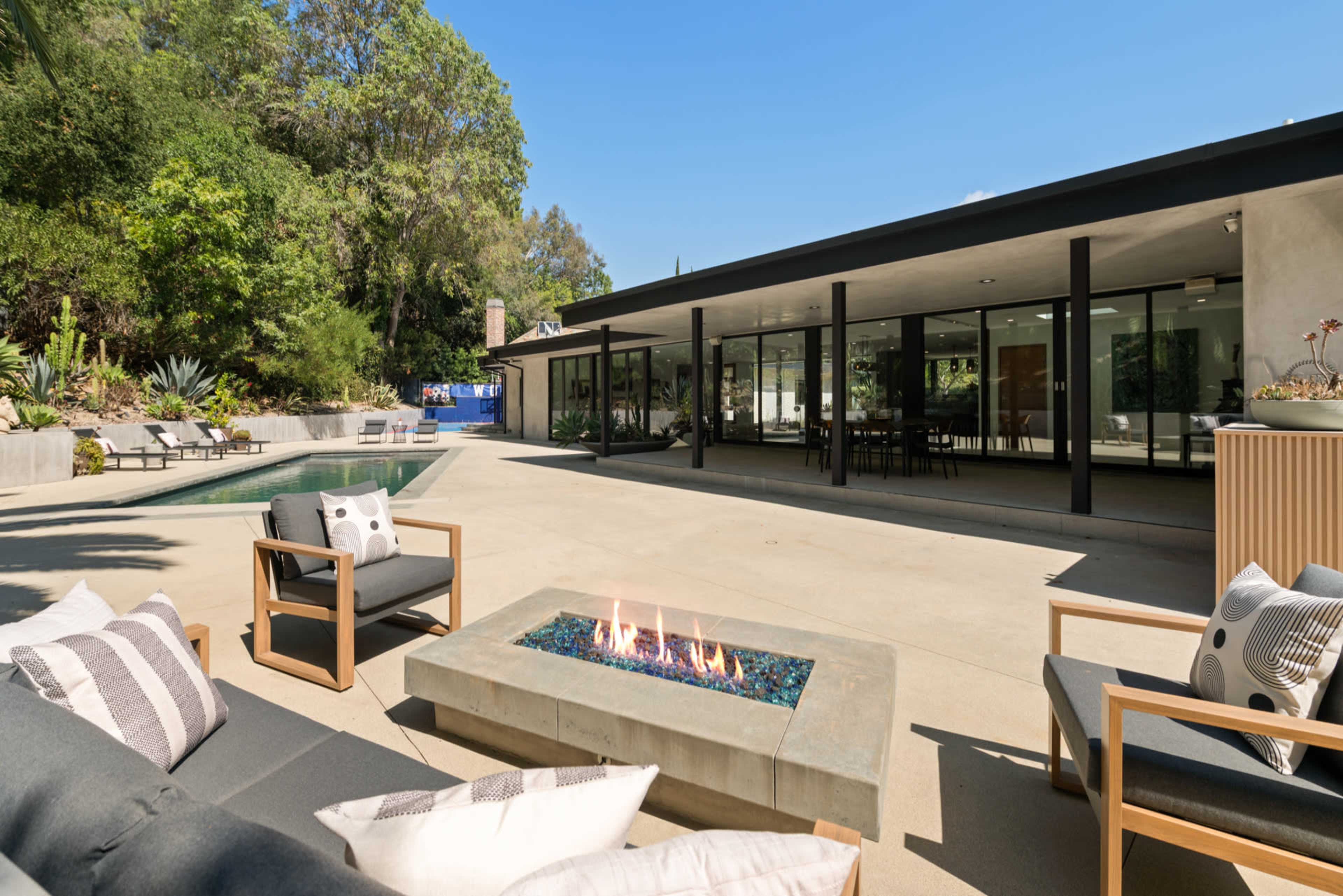 A modern outdoor living space features a fire pit surrounded by seating, with a pool and large glass windows of a house in the background, set against a lush green landscape.