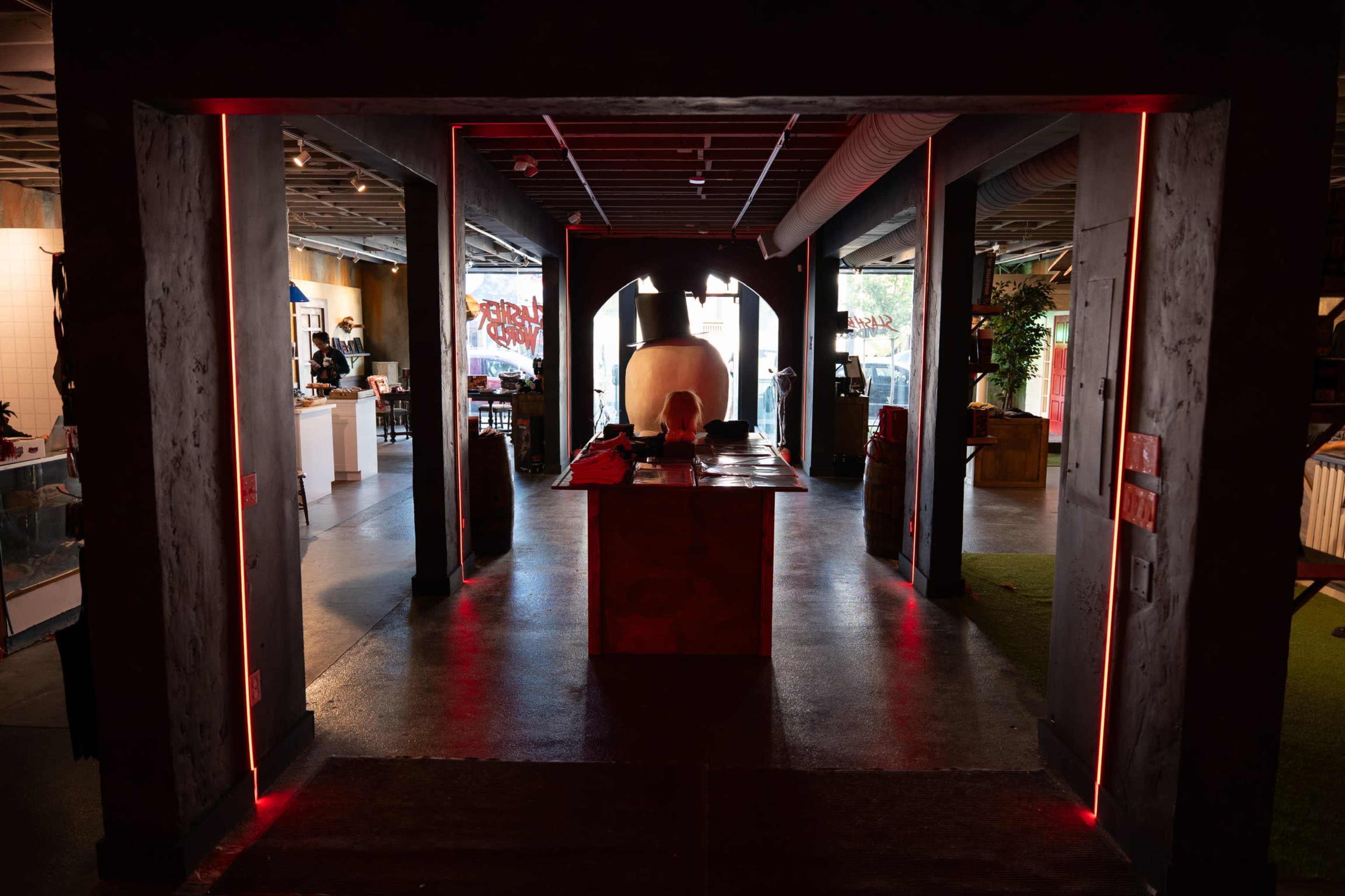 The image shows a dimly lit entryway leading into a modern space with red accent lighting, featuring a large, abstract sculpture in the center.