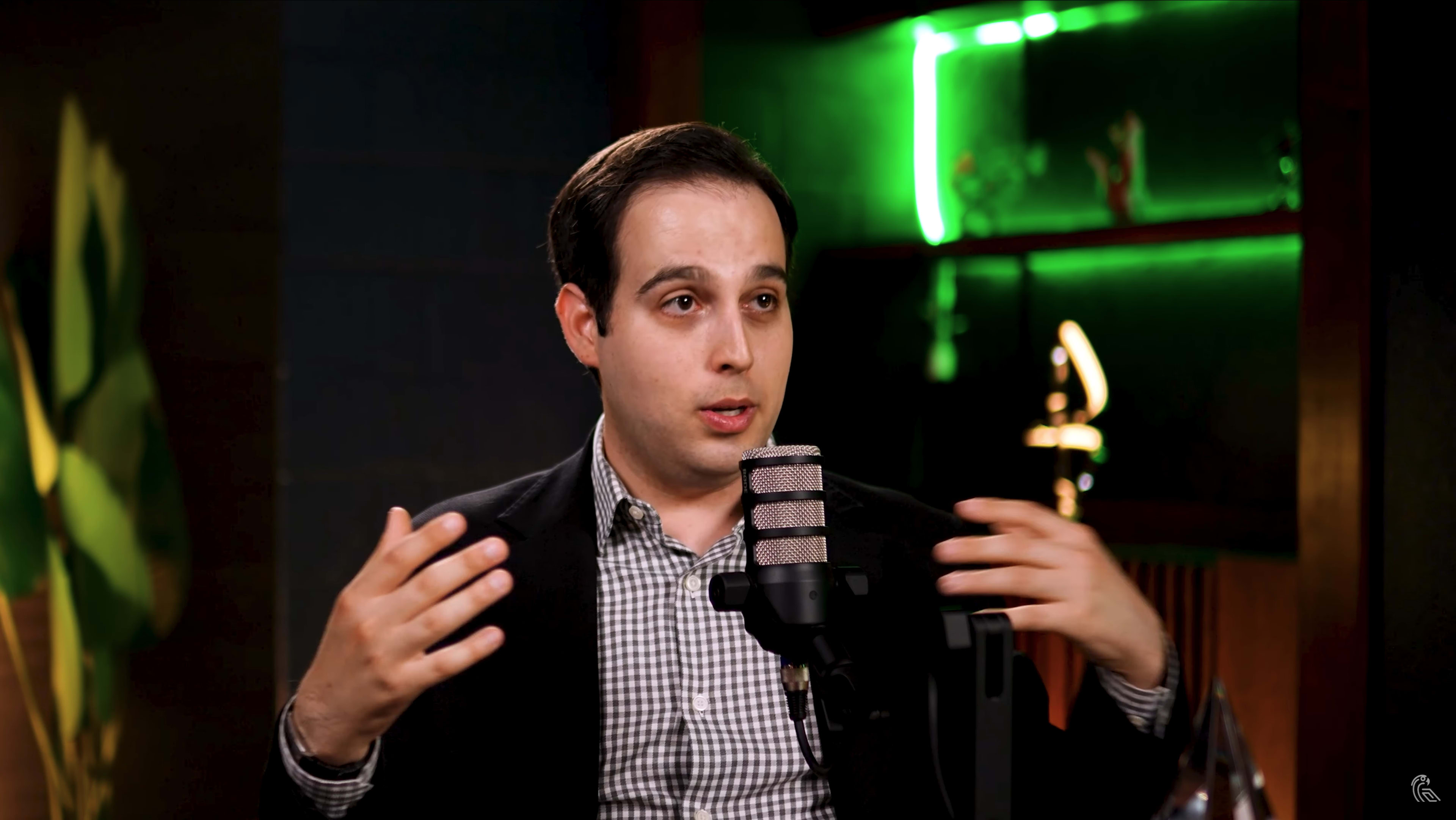 A man in a black blazer gestures while speaking into a microphone in a well-lit room with shelves displaying decorative items.