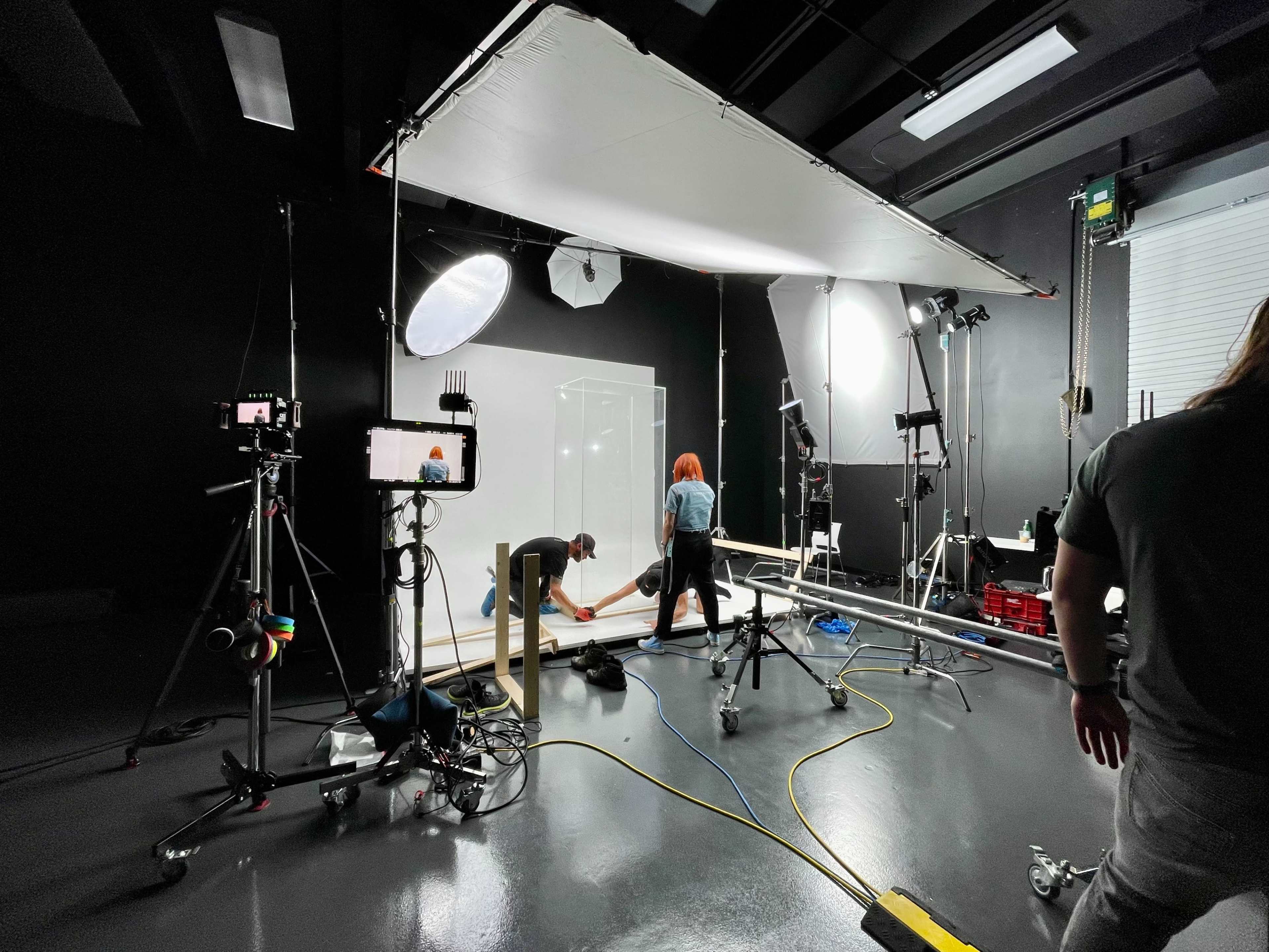 A film crew sets up equipment in a dark studio with various lights and cameras for a shoot.