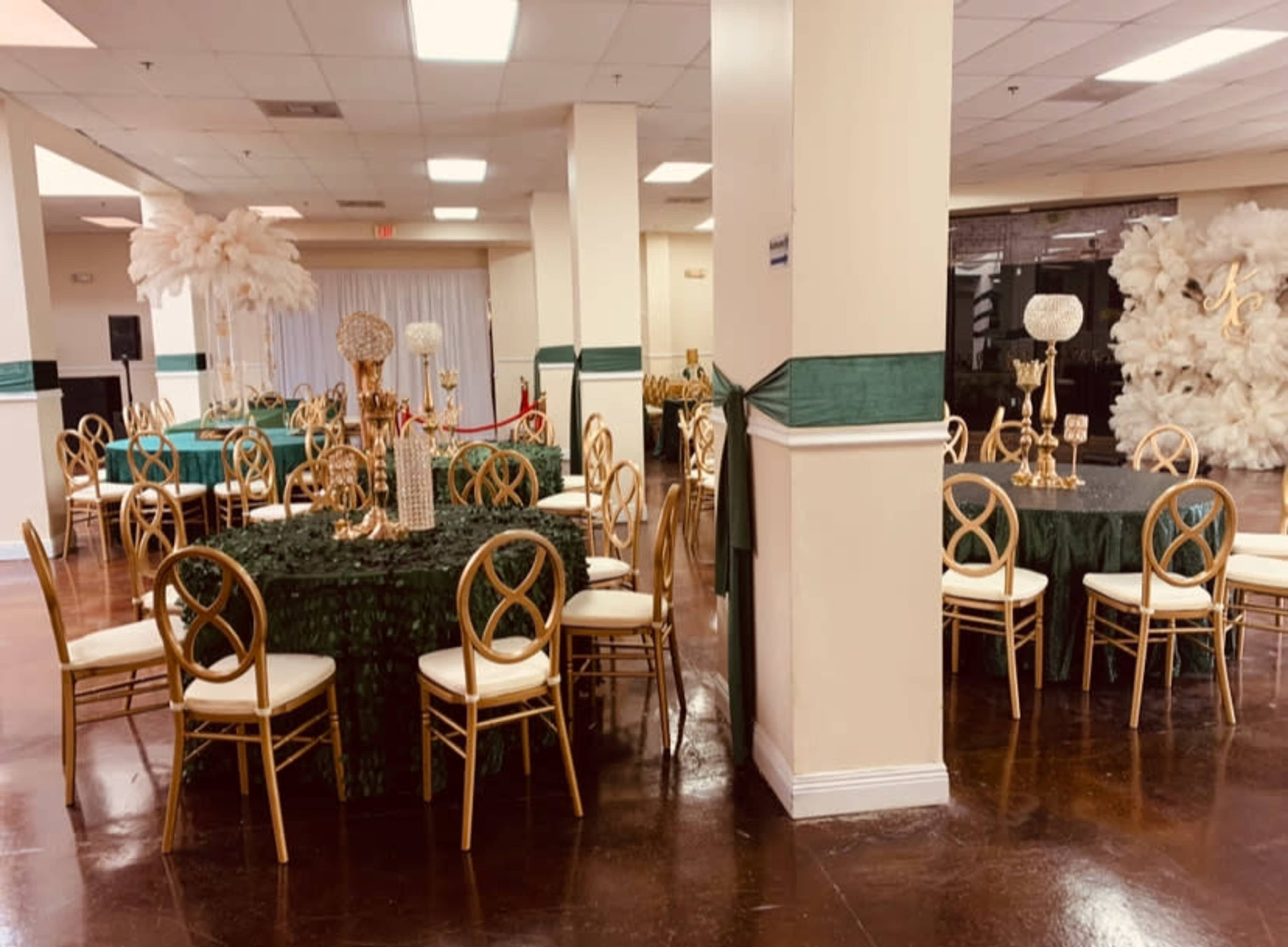 The image shows a banquet hall set up for an event, featuring round tables with green and gold table settings and decorative centerpieces, surrounded by gold chairs.