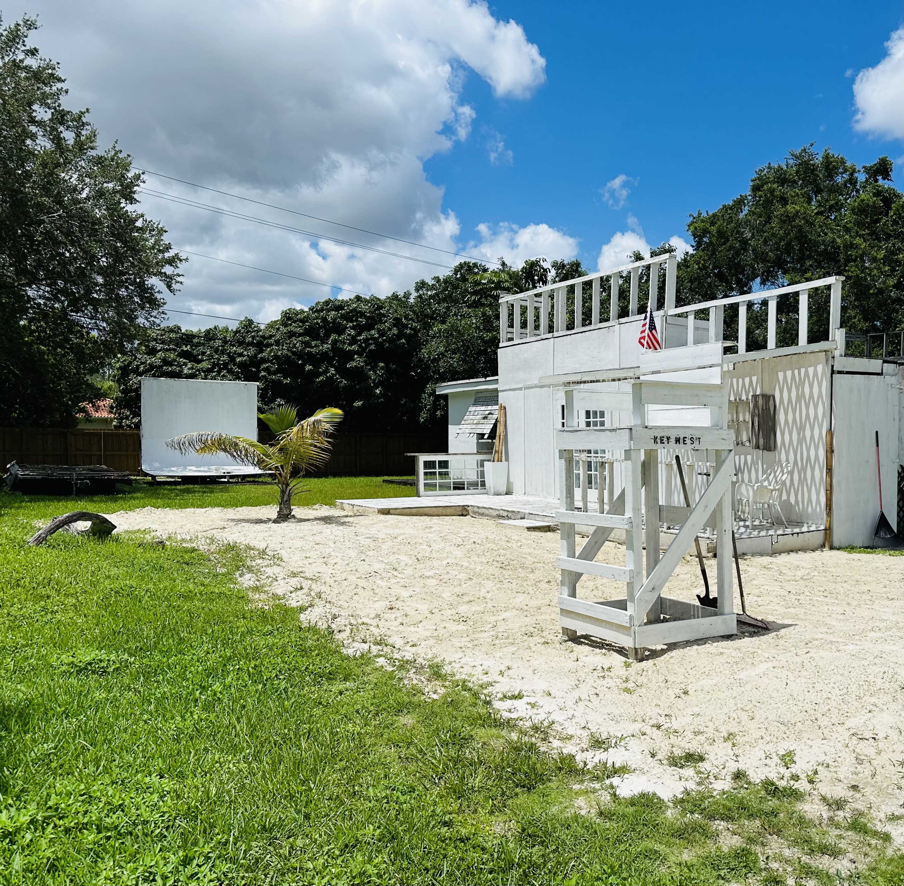The image shows a white building with a lifeguard stand and a palm tree in a sandy area, surrounded by grass and trees under a partly cloudy sky.
