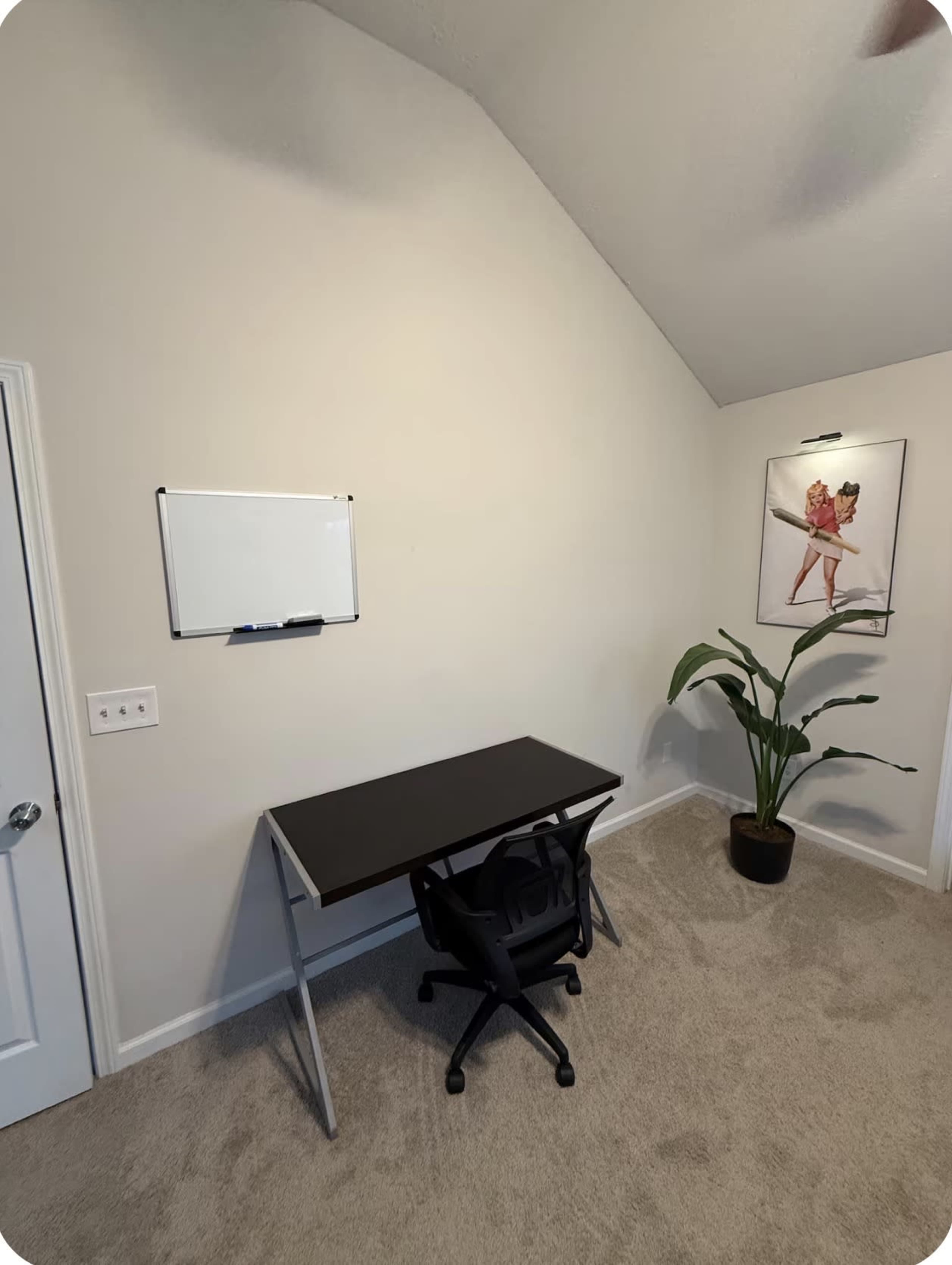 The image shows a simple home office setup with a black desk, an office chair, a whiteboard on the wall, and a potted plant in the corner.