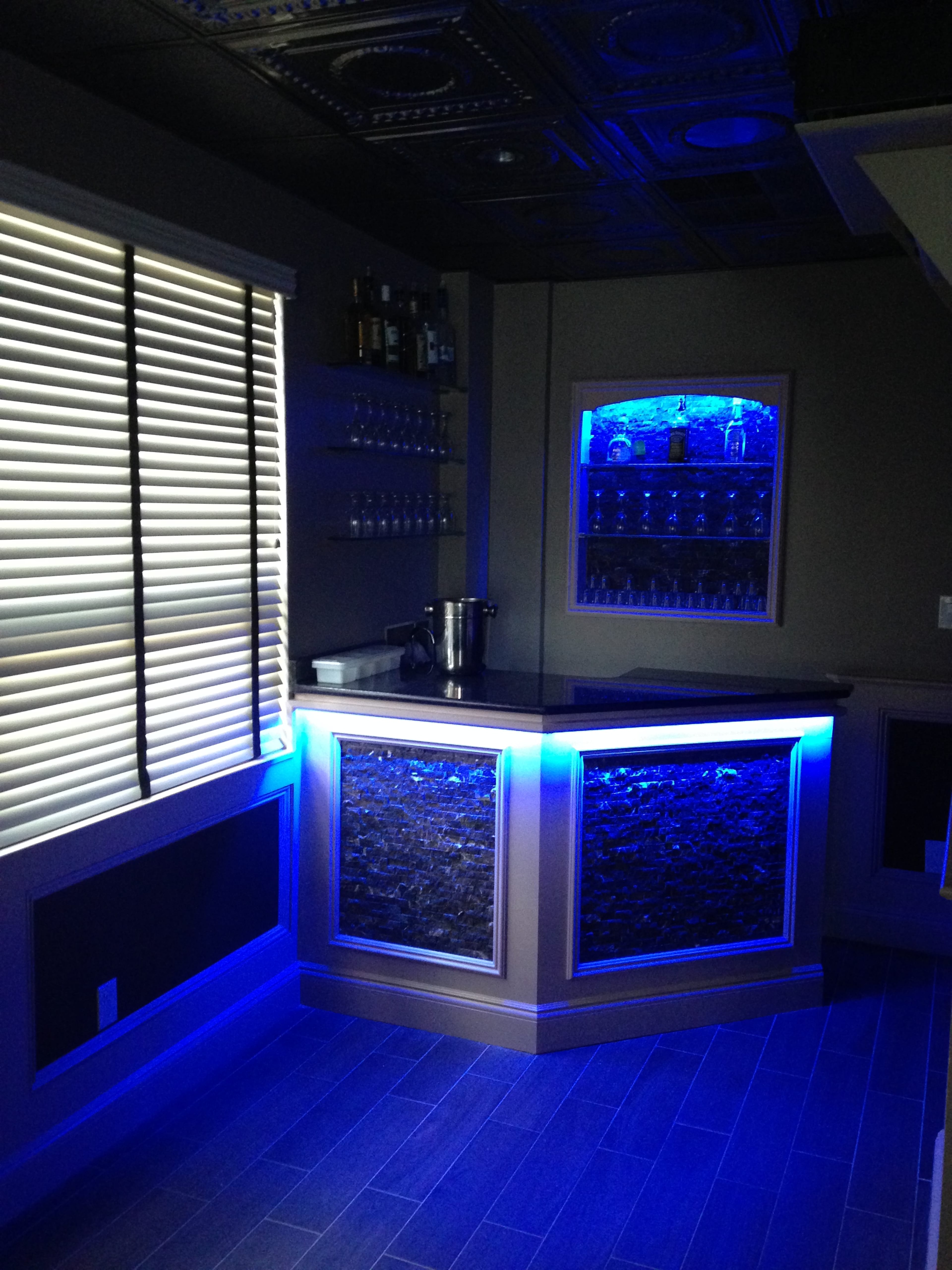 A modern home bar with illuminated blue accents, a countertop, and shelves displaying various bottles.