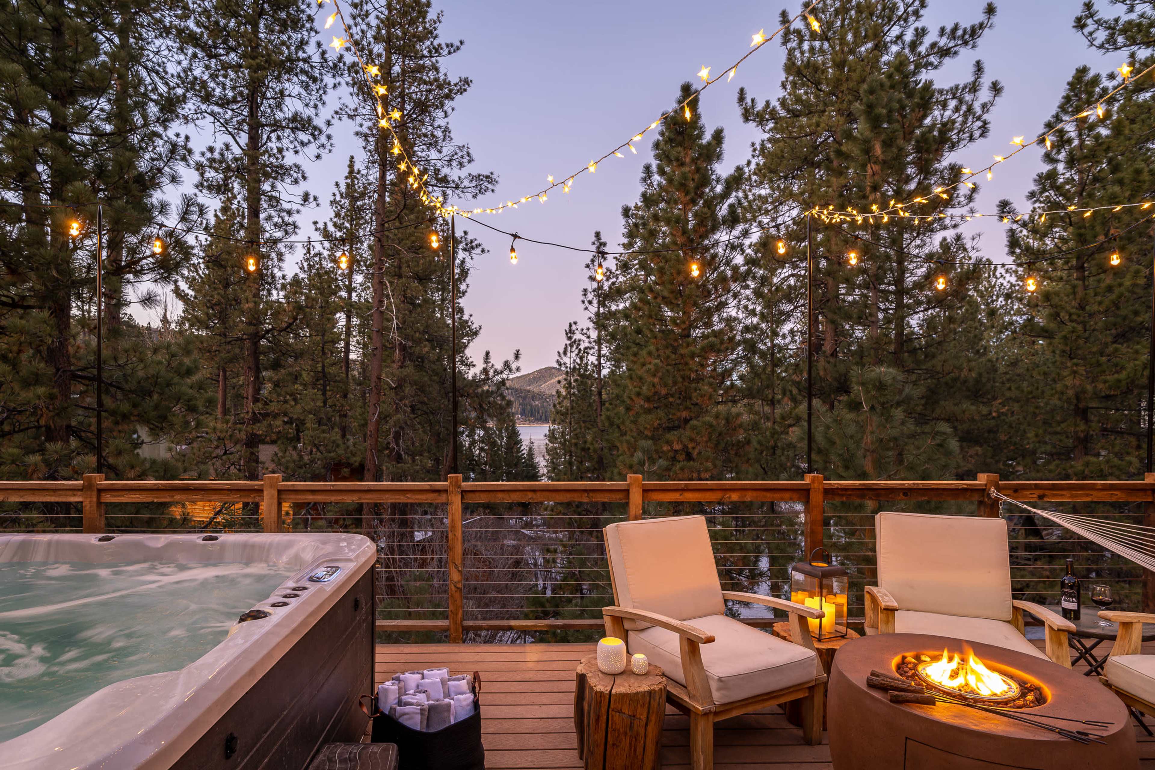 A hot tub and two chairs are positioned on a wooden deck surrounded by tall trees and twinkling lights, overlooking a body of water at dusk.