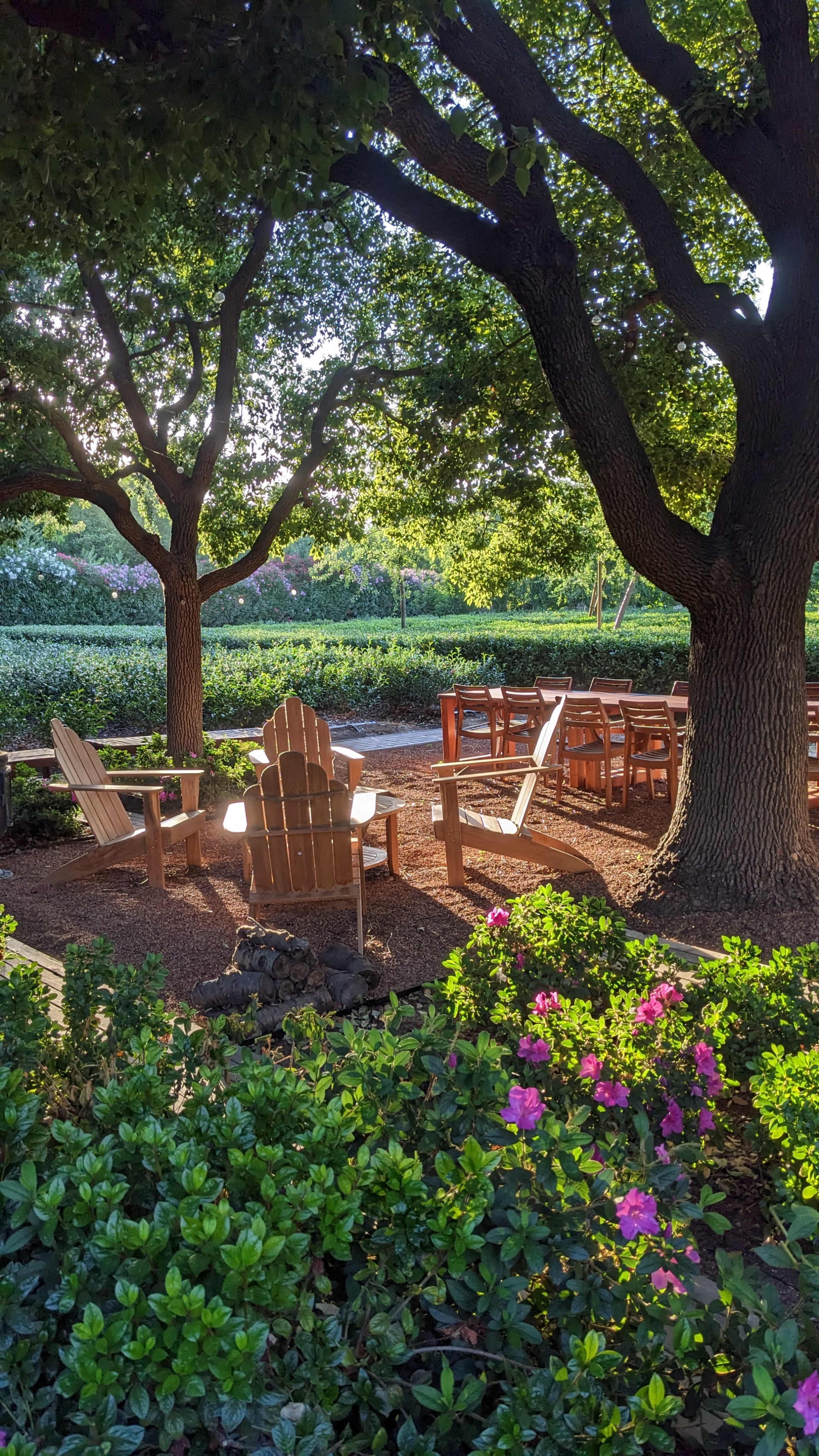 The scene features wooden Adirondack chairs arranged in a garden area surrounded by lush greenery and blooming flowers.