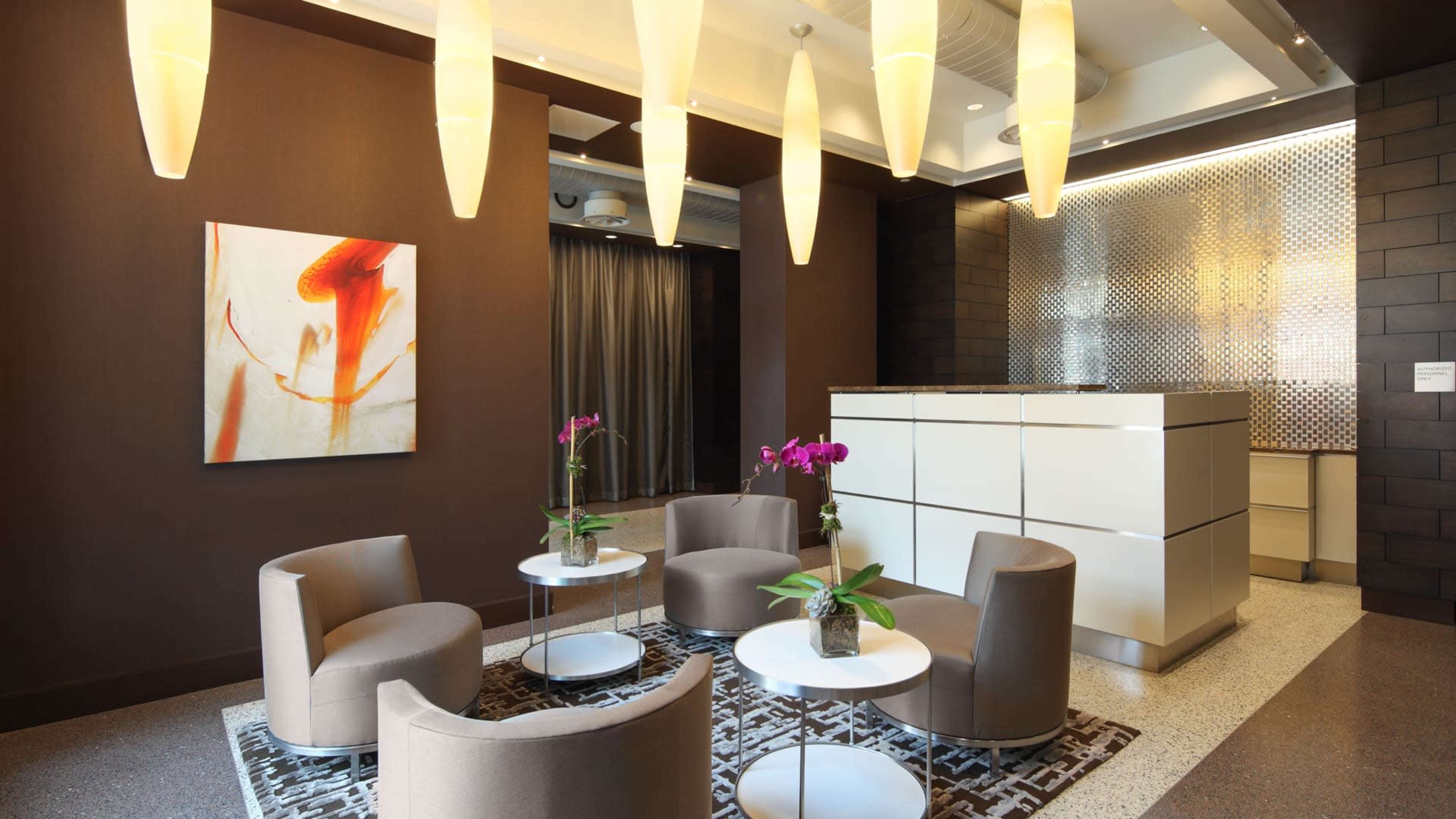 The image shows a modern lobby area featuring a reception desk, soft seating arrangements with round tables, and decorative lighting.