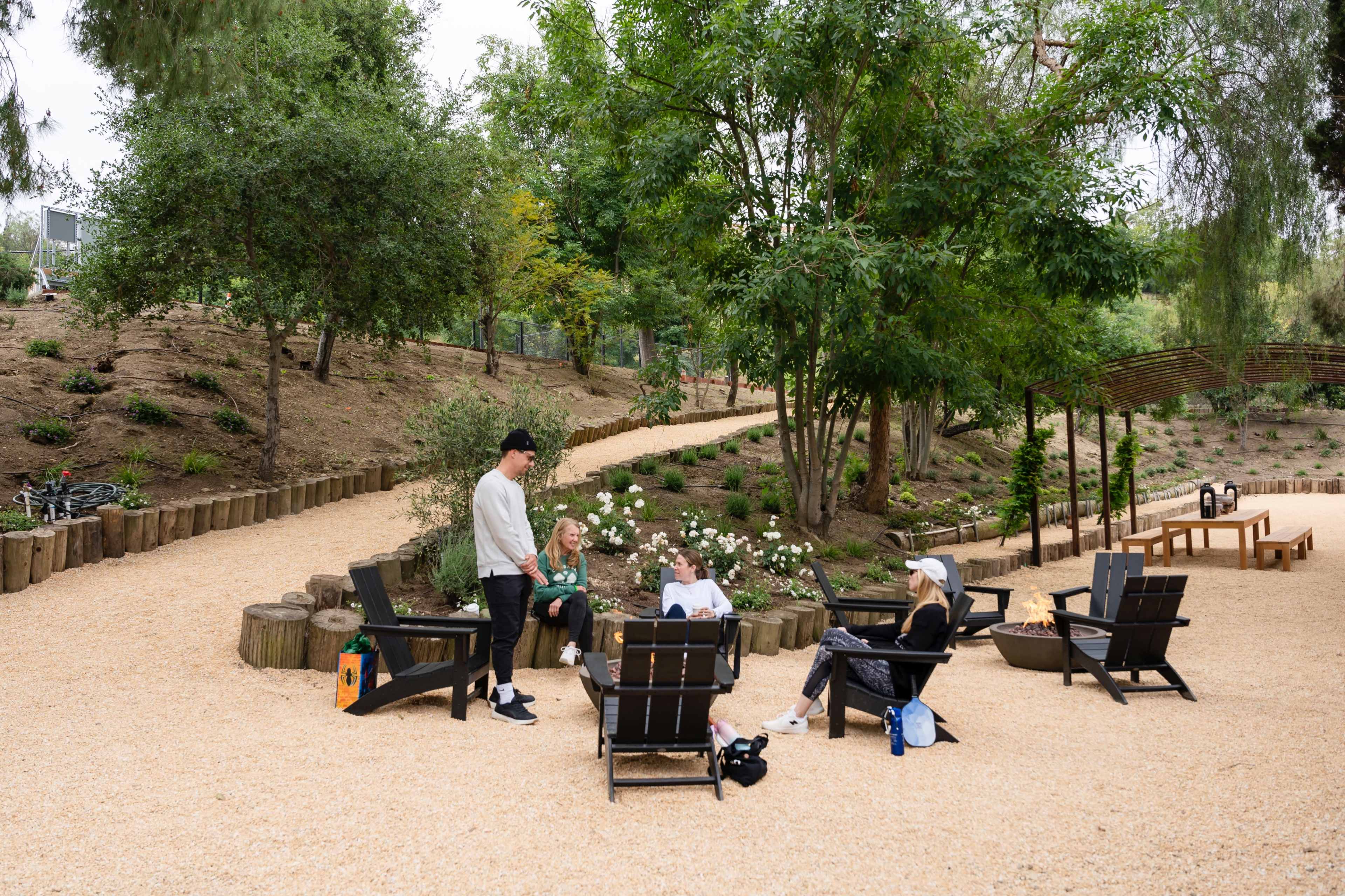 A group of four people gathers around Adirondack chairs in a landscaped area with a gravel path and greenery.