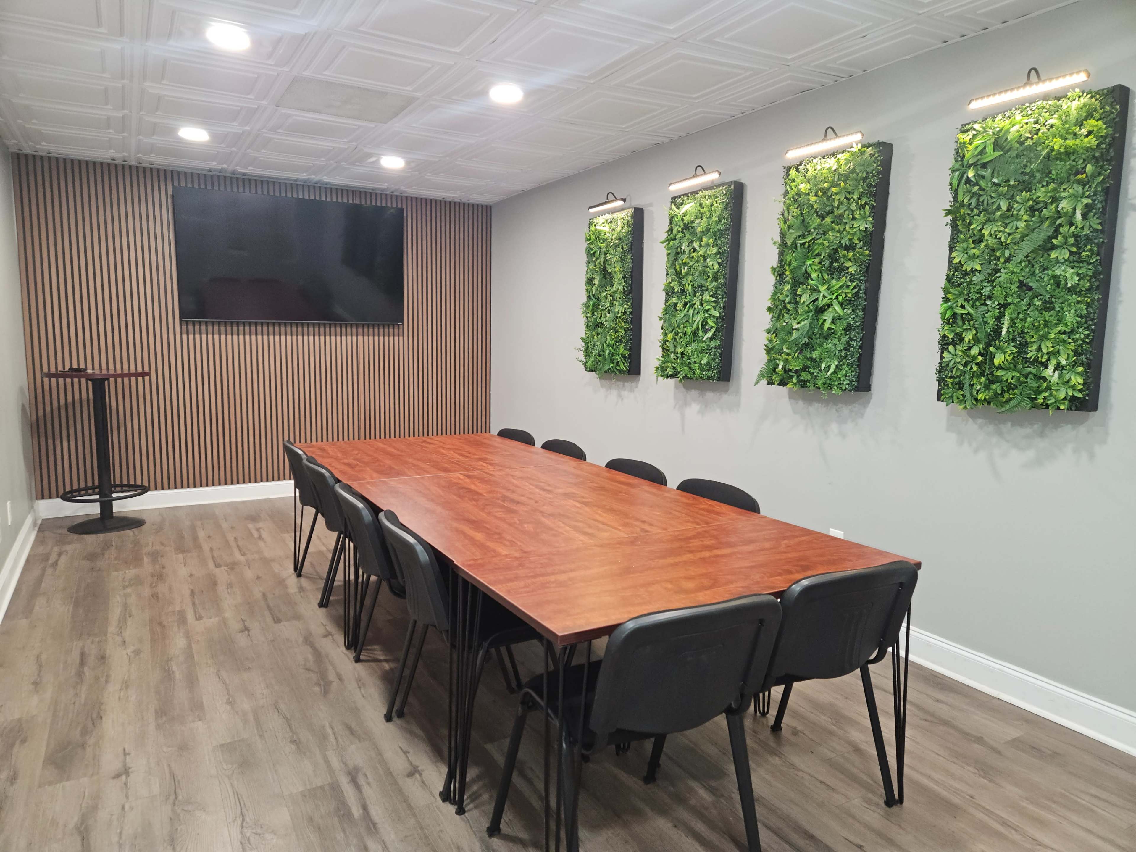 The image shows a modern conference room featuring a long wooden table surrounded by black chairs, with green vertical planters on the wall and a television mounted above.