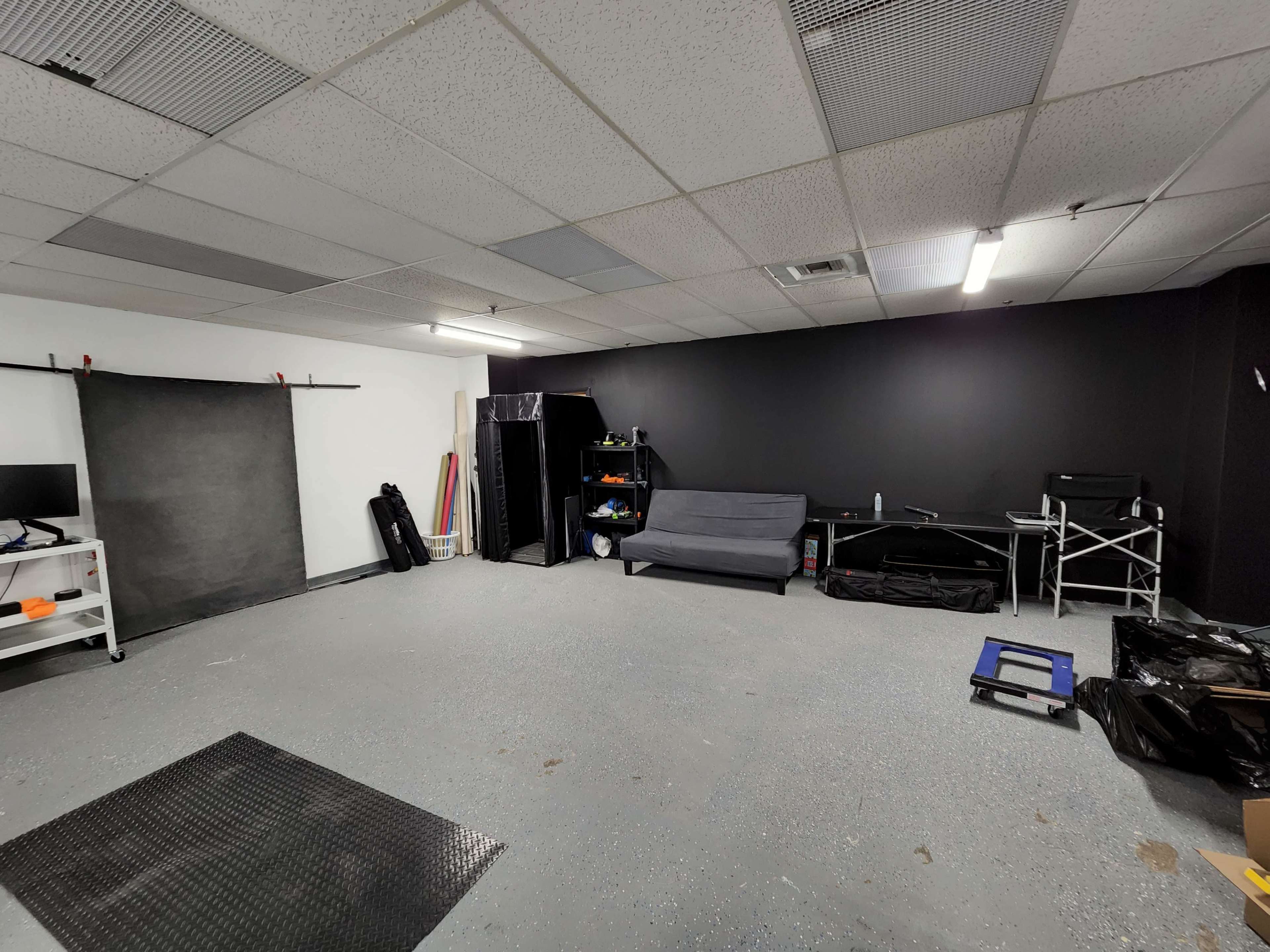 A spacious, empty room with a gray concrete floor, a black wall, and minimal furniture, including a gray couch and a few storage items.