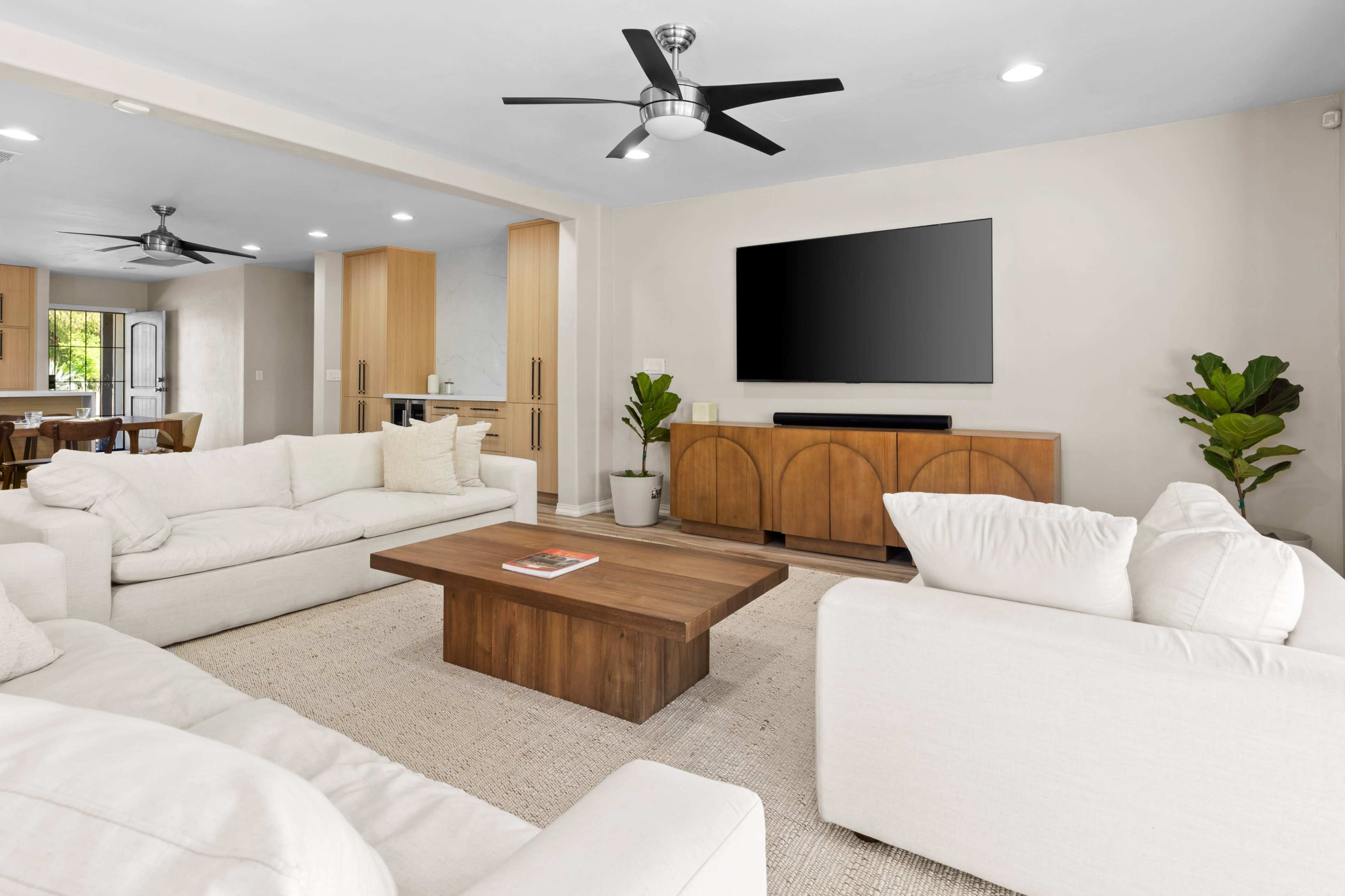 The image shows a modern living room with two white sofas facing a wooden coffee table and a wall-mounted television, surrounded by neutral-colored walls and a plant in the corner.