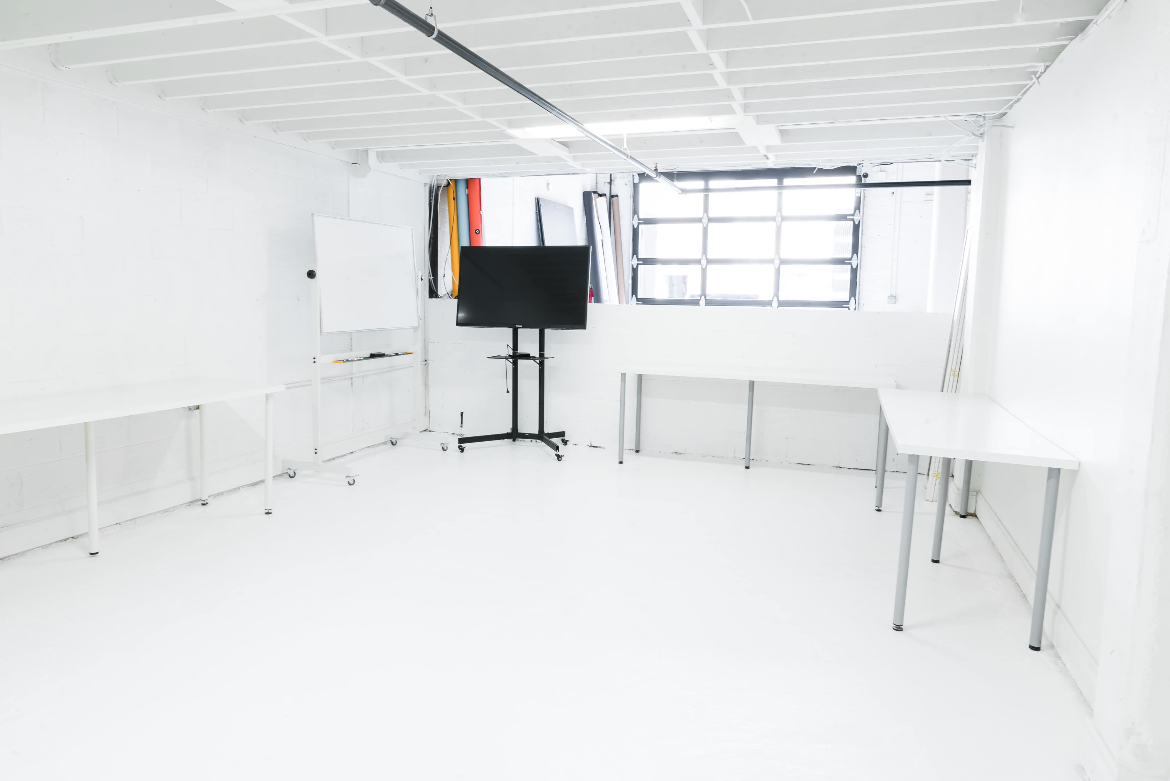 The image shows a spacious, white-walled room with two tables, a large monitor on a mobile stand, and a whiteboard.