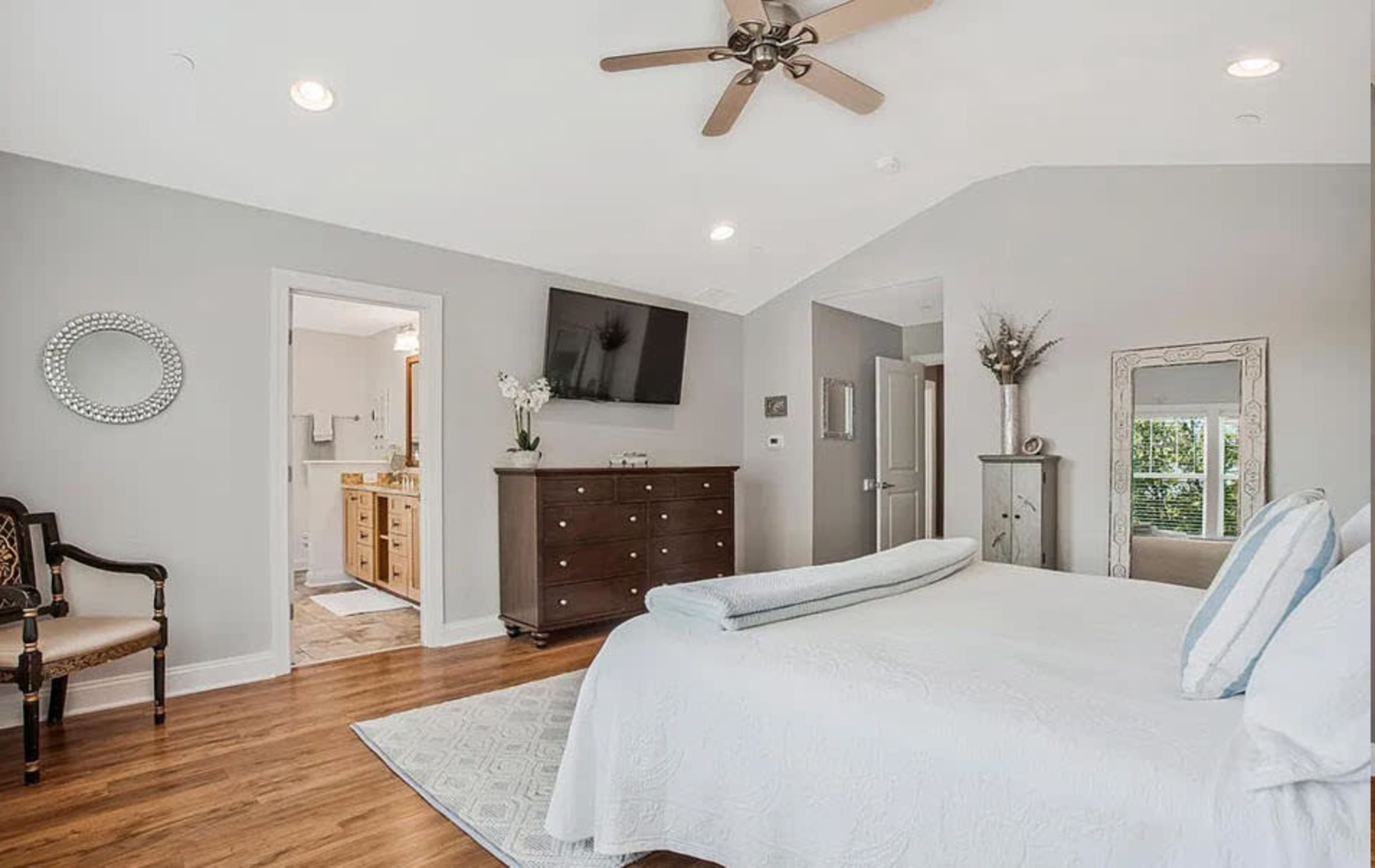 The image shows a spacious, well-lit bedroom featuring a large bed, a dresser, a television mounted on the wall, and an adjoining bathroom visible through an open door.