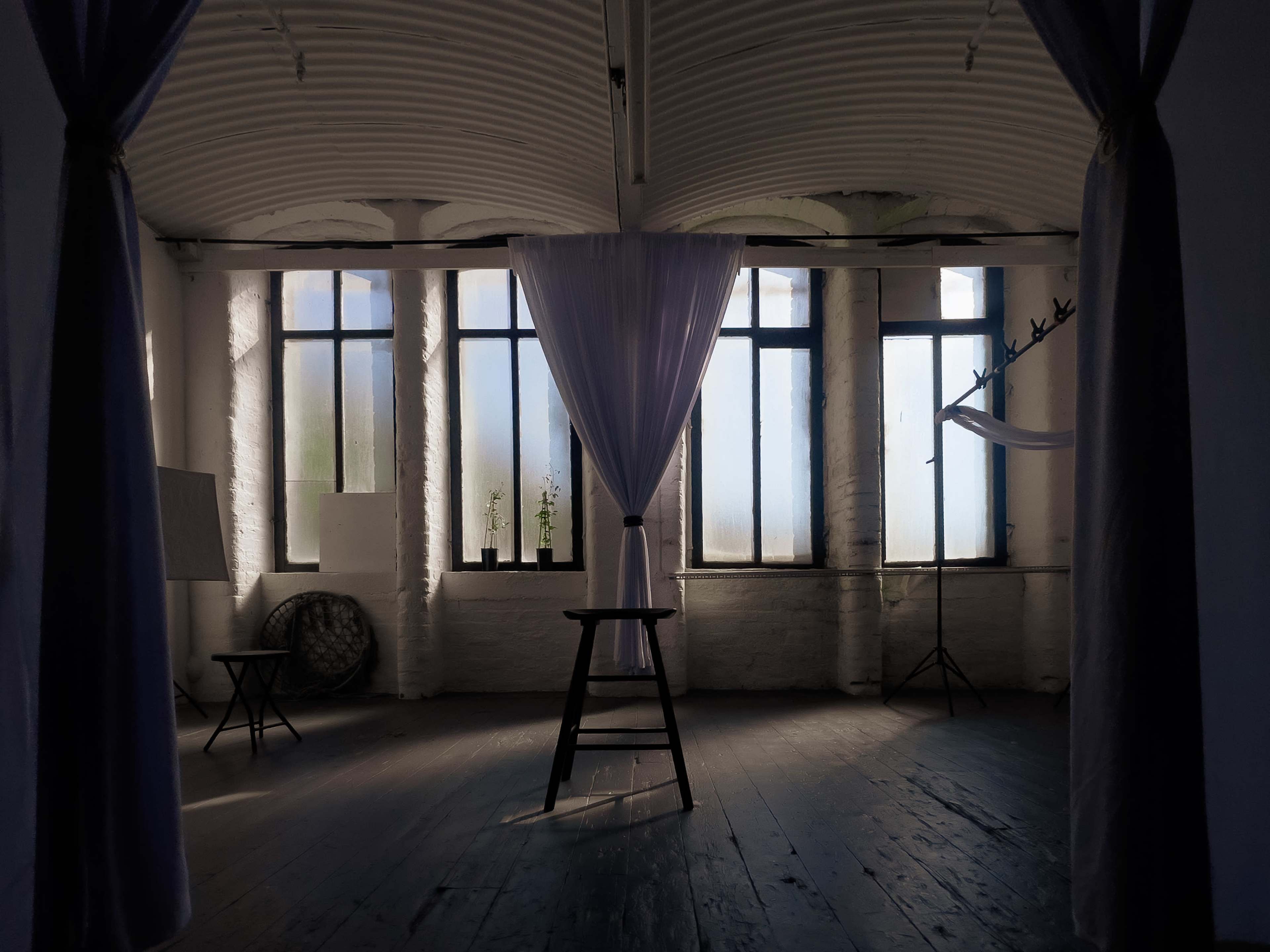 The image shows a dimly lit room featuring tall windows, a standing stool, a curtain, and minimalist furnishings.