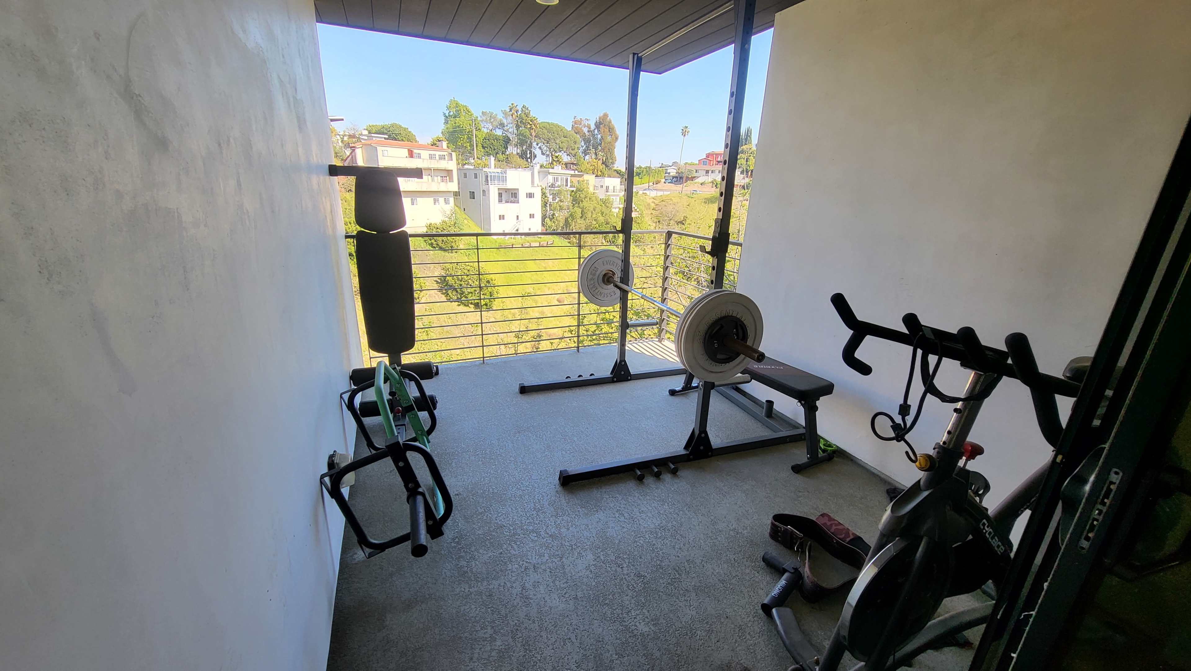 The image shows a small gym area on a balcony with an exercise bike, weights, and a rowing machine, overlooking a green hillside.