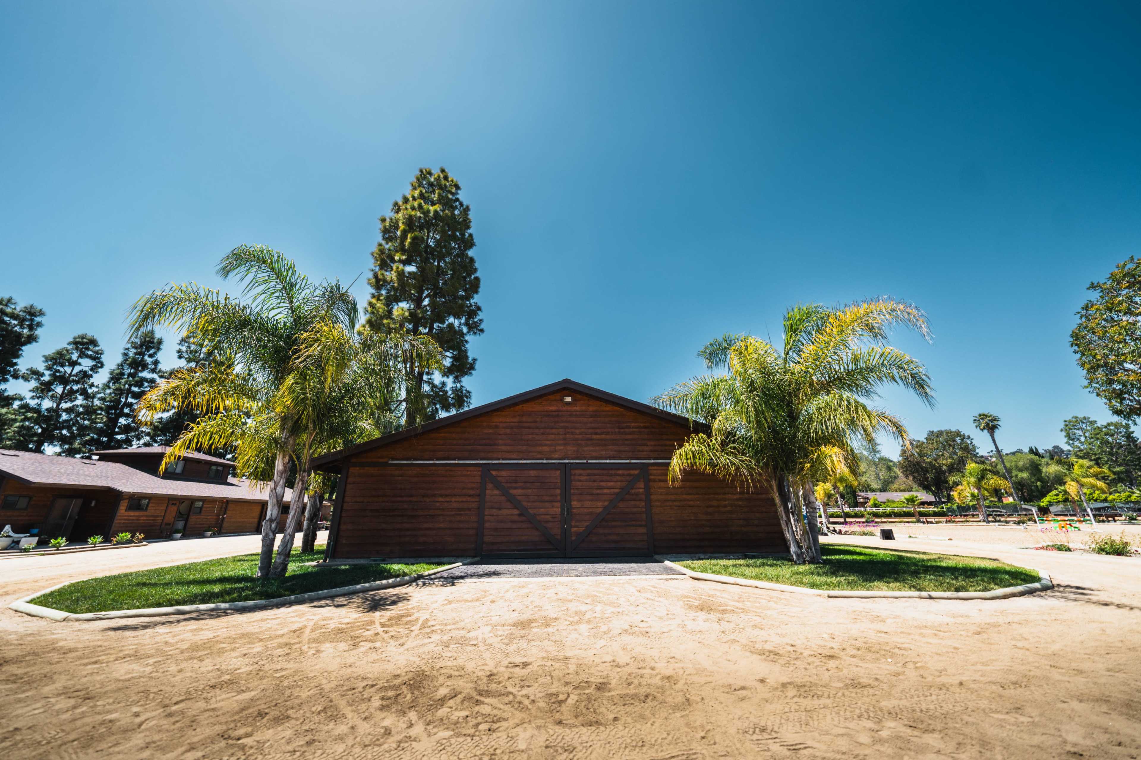 A wooden barn with palm trees is set against a clear blue sky in a landscaped area.