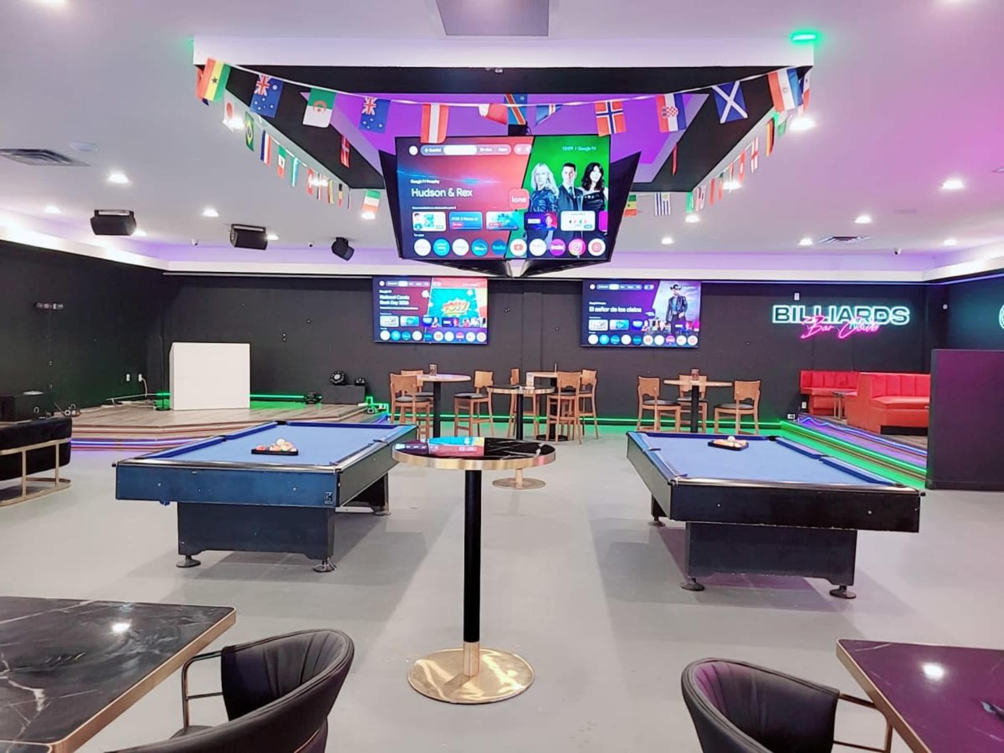 A modern billiards room features two pool tables, seating areas, and multiple screens displaying sports and game information, with flags from various countries hanging on the walls.