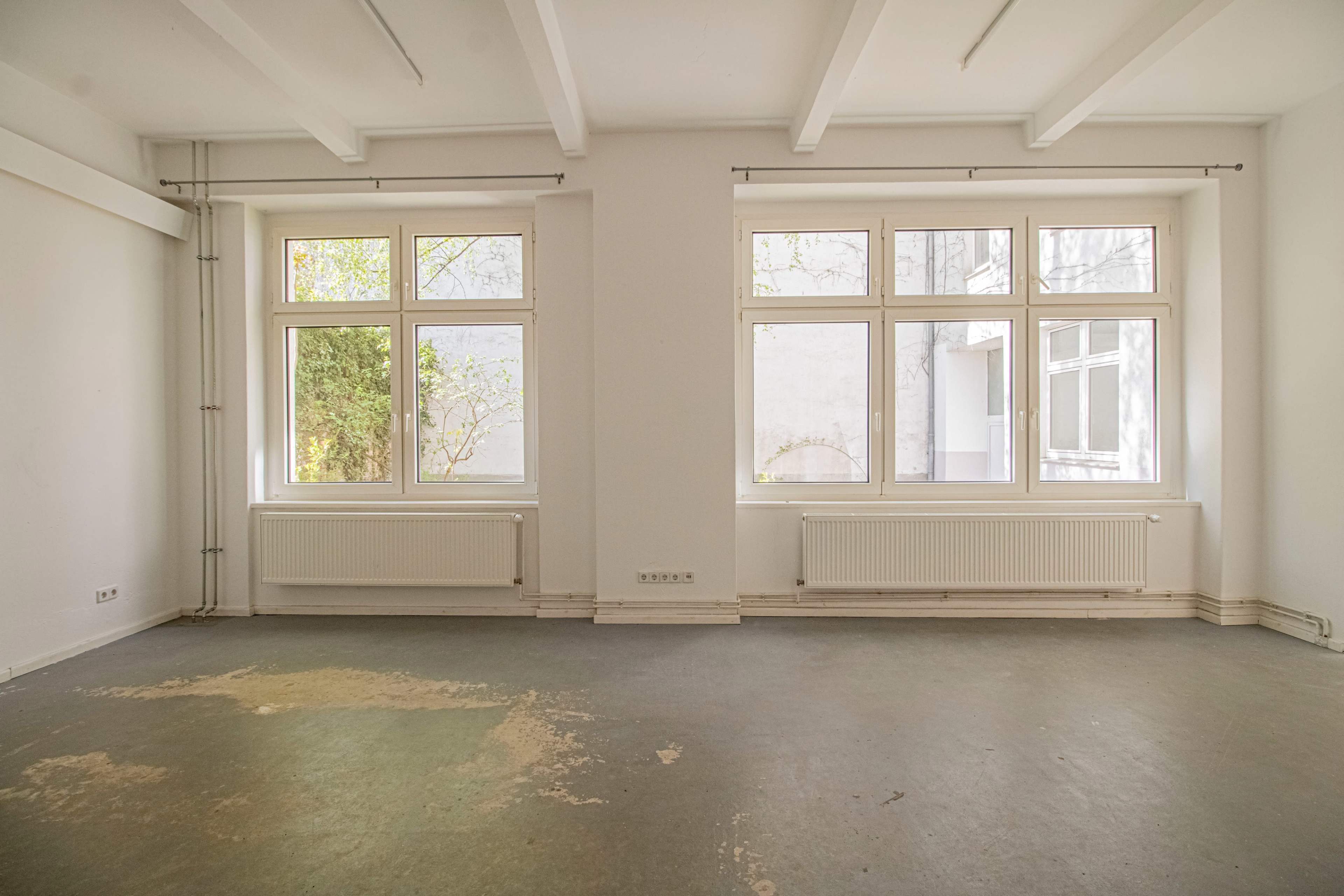 The image shows a spacious, empty room with two large windows and a bare concrete floor.