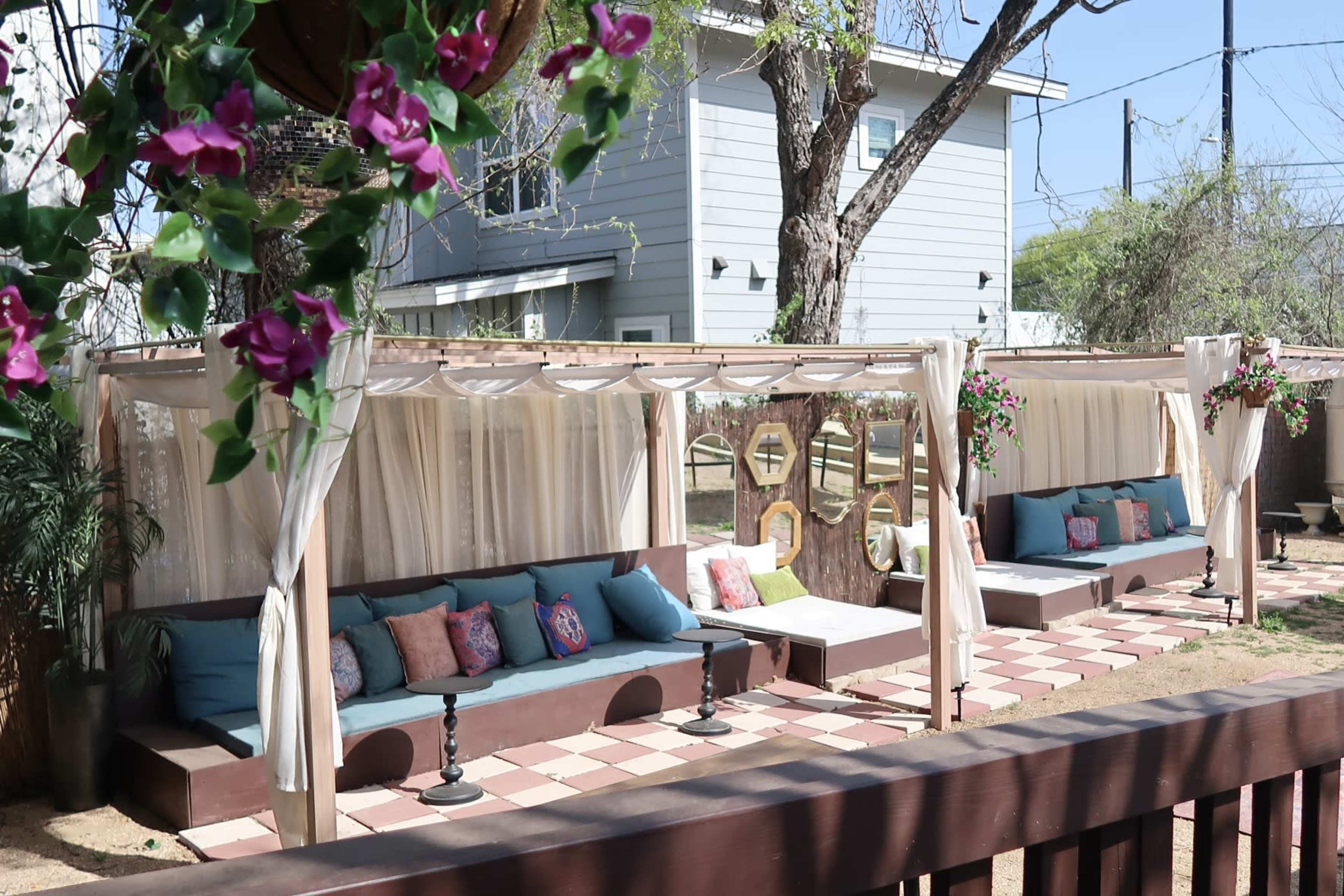 The image features a shaded outdoor seating area with upholstered benches and decorative pillows, surrounded by greenery and a wooden fence.