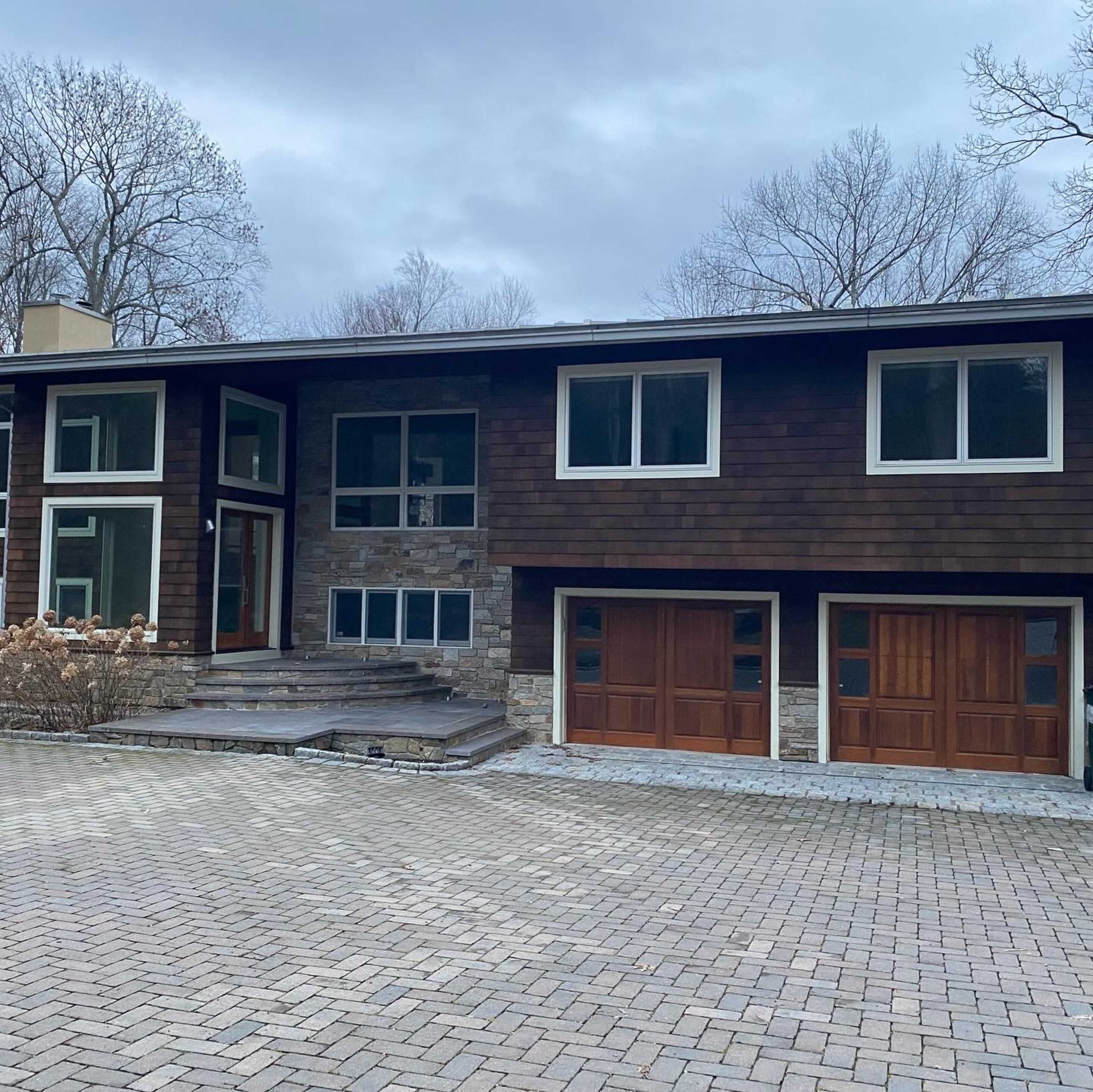 The image shows a modern two-story house with a combination of wood and stone exterior, featuring large windows and two garage doors.