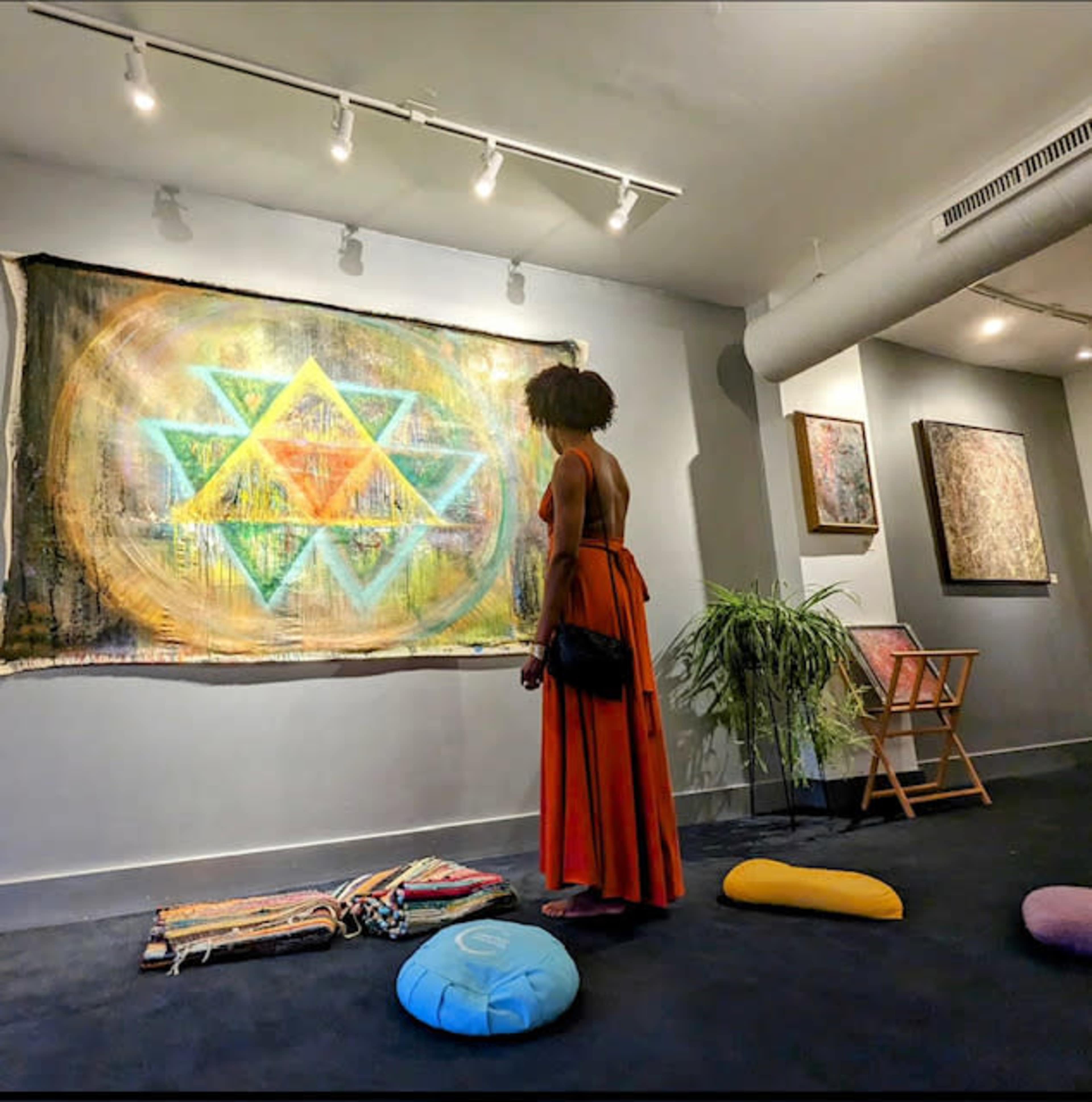 A woman in an orange dress stands in an art gallery, gazing at a colorful geometric tapestry on the wall.