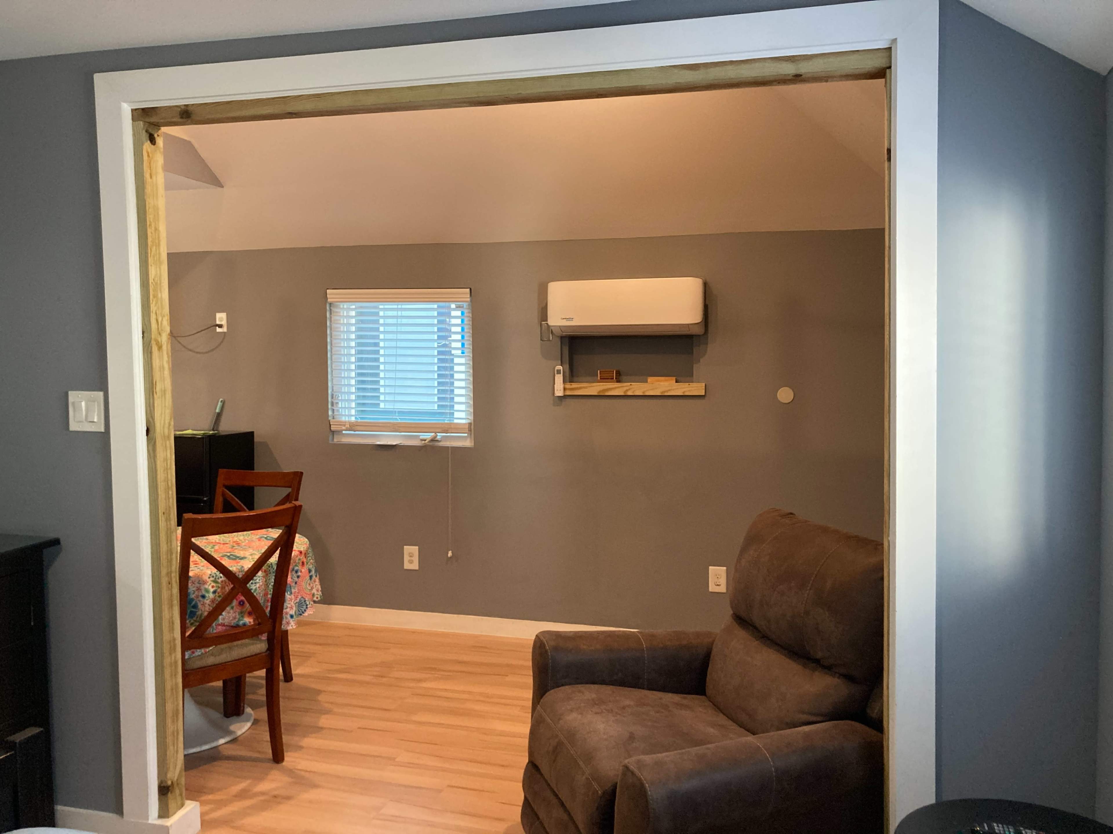 An open doorway leading from one room to another, with a brown armchair, a dining table with a patterned tablecloth, and a wall-mounted air conditioner visible in the adjacent room.