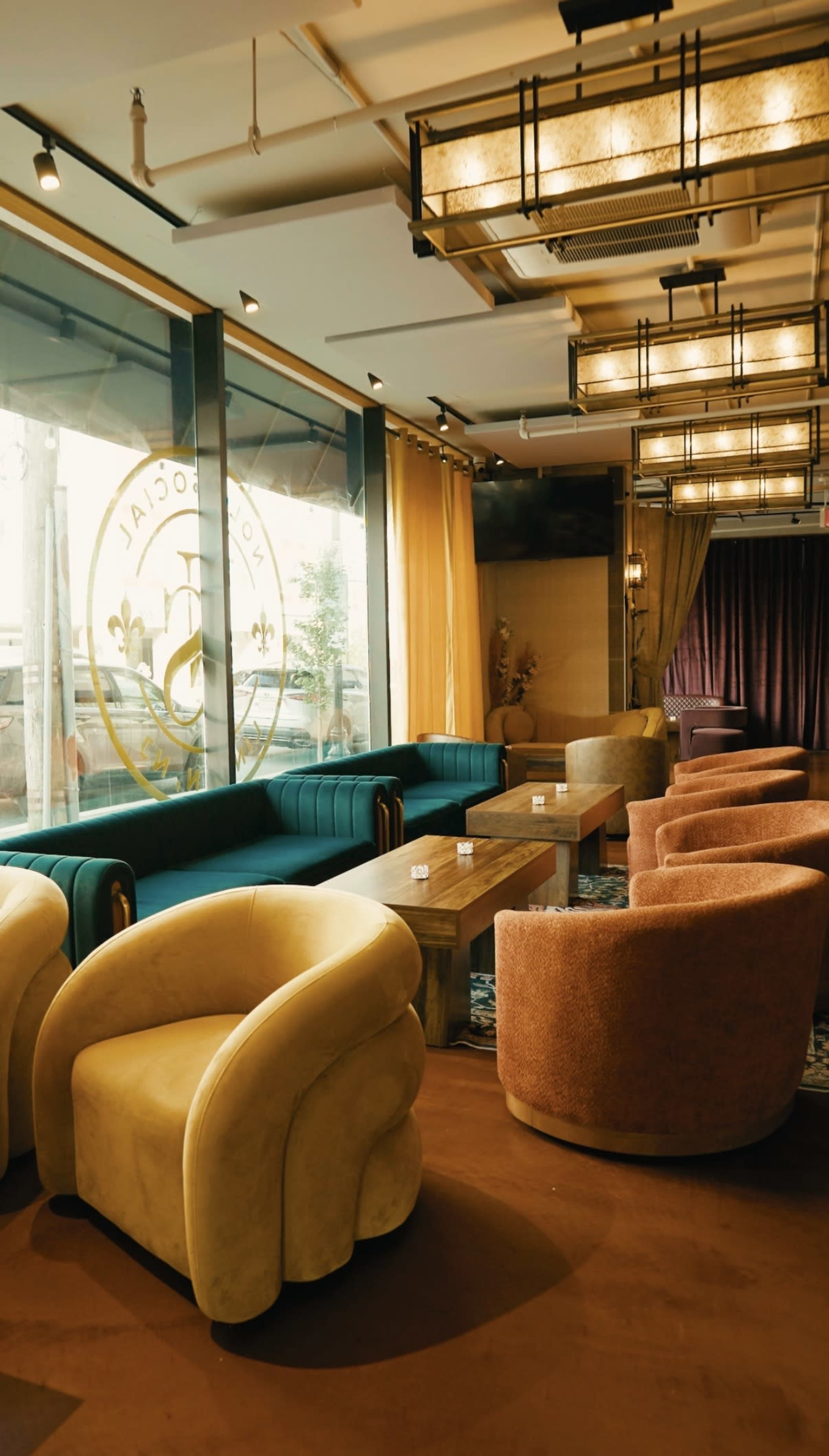 The image shows a modern lounge area with green and orange upholstered chairs, wooden tables, and large windows allowing natural light.