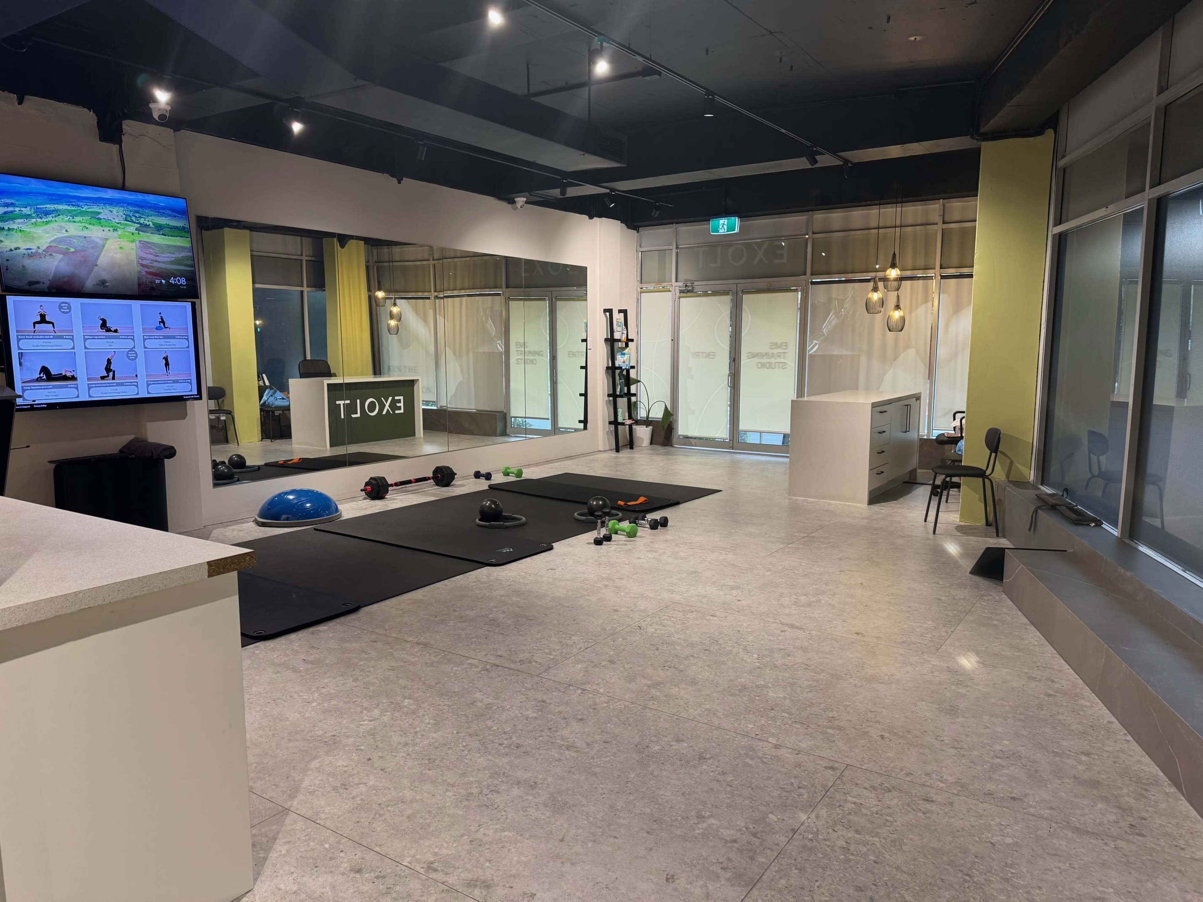 The image shows a modern fitness studio with exercise equipment, including dumbbells, mats, and a large screen displaying workout information.