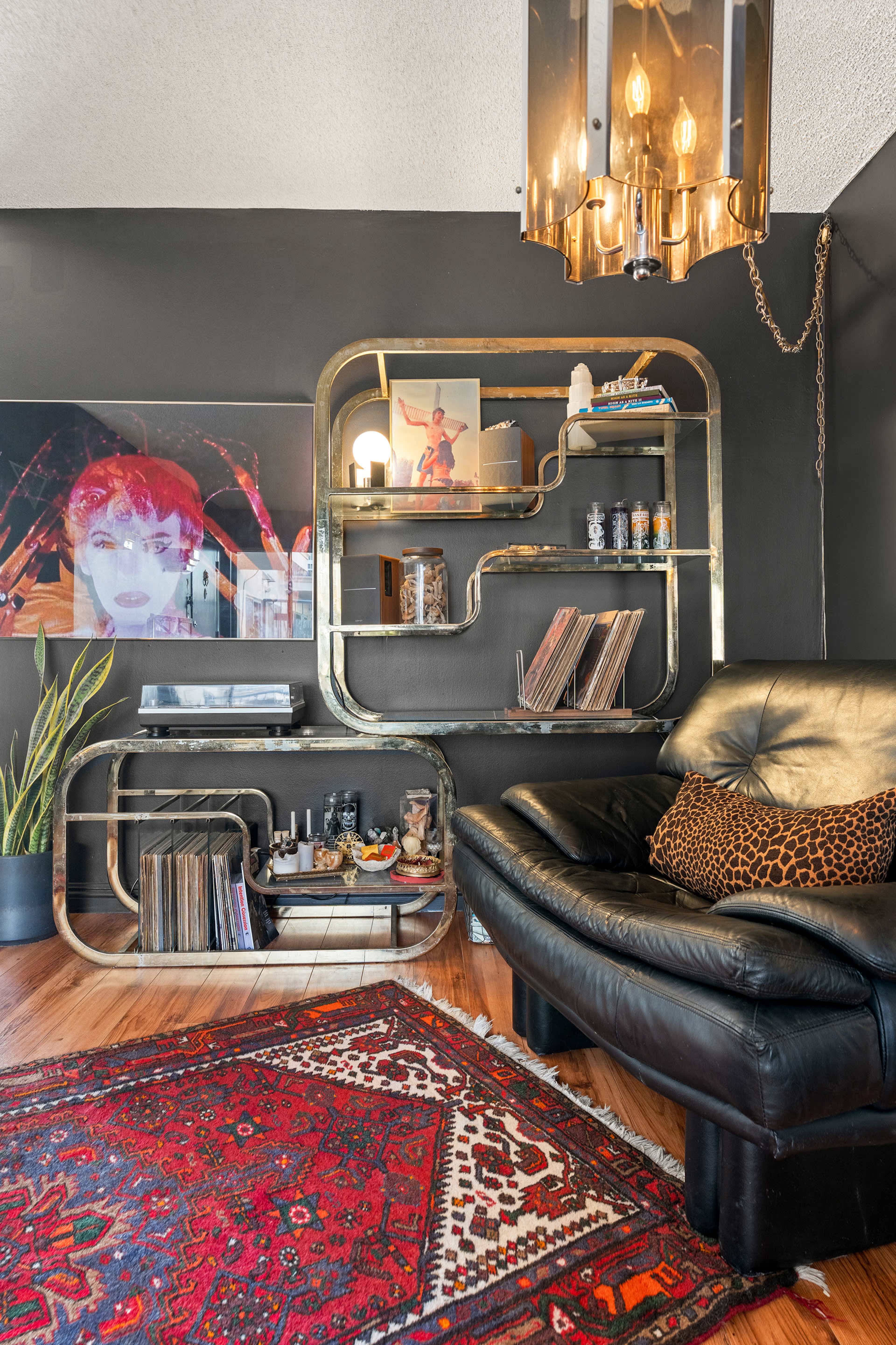 The image shows a modern living room featuring a black leather chair, a stylish bookshelf with decorative items, a turntable and vinyl records, and a colorful area rug on a hardwood floor.