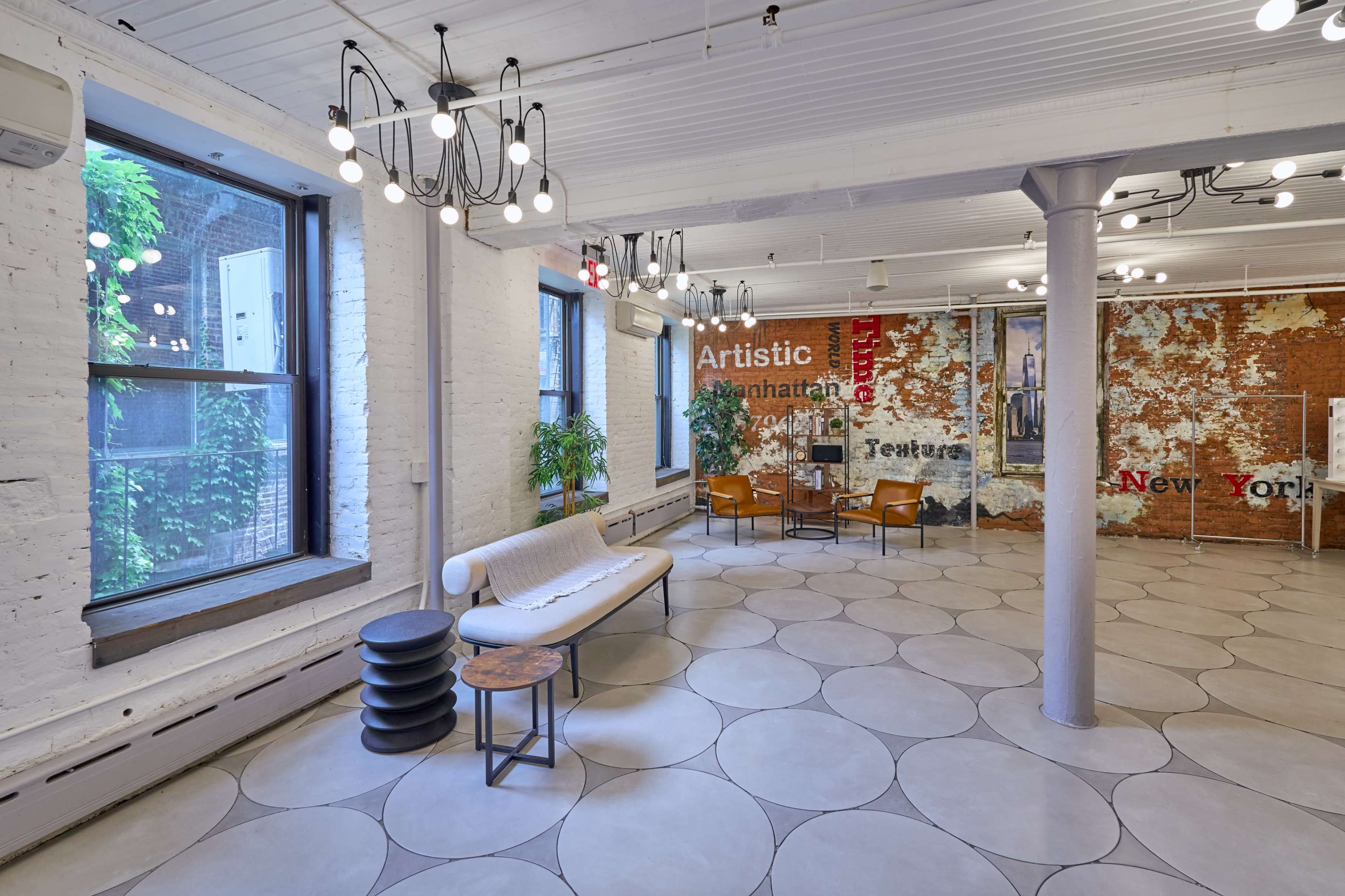 The image shows a spacious, contemporary room with white brick walls, large windows, and a circular tile floor, featuring a seating area with a sofa and chairs.