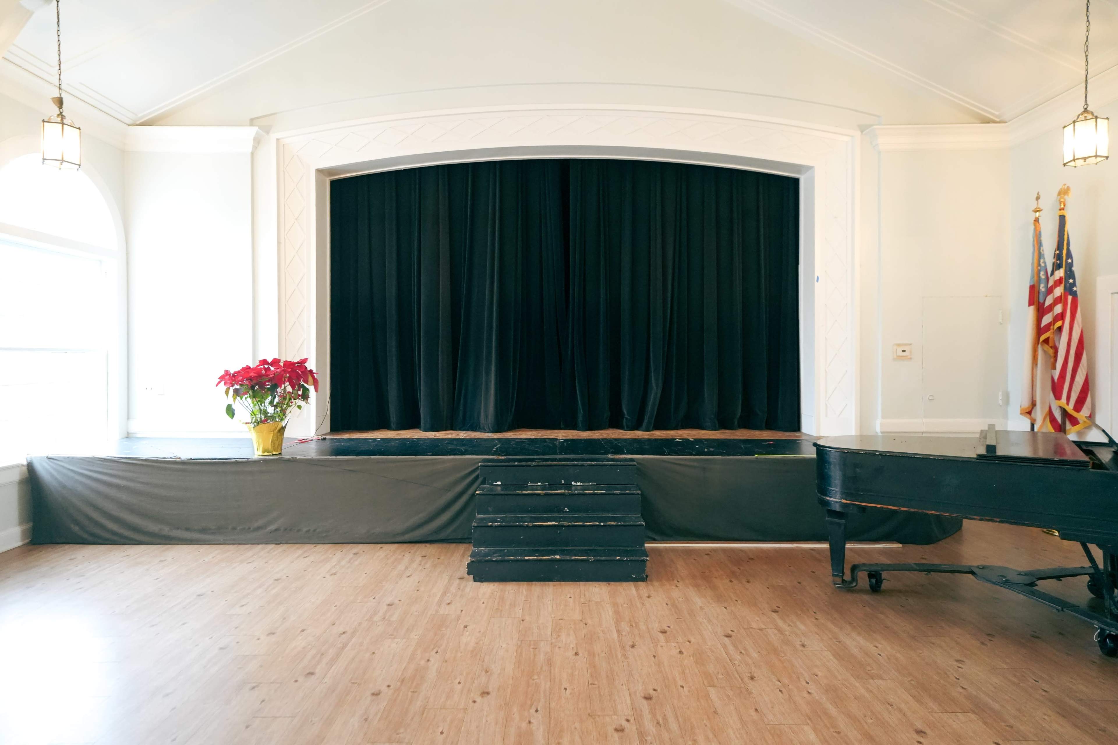 The image features a stage with black curtains, a wooden floor, a few steps leading up to the stage, a potted plant in the foreground, and an upright piano on the side.