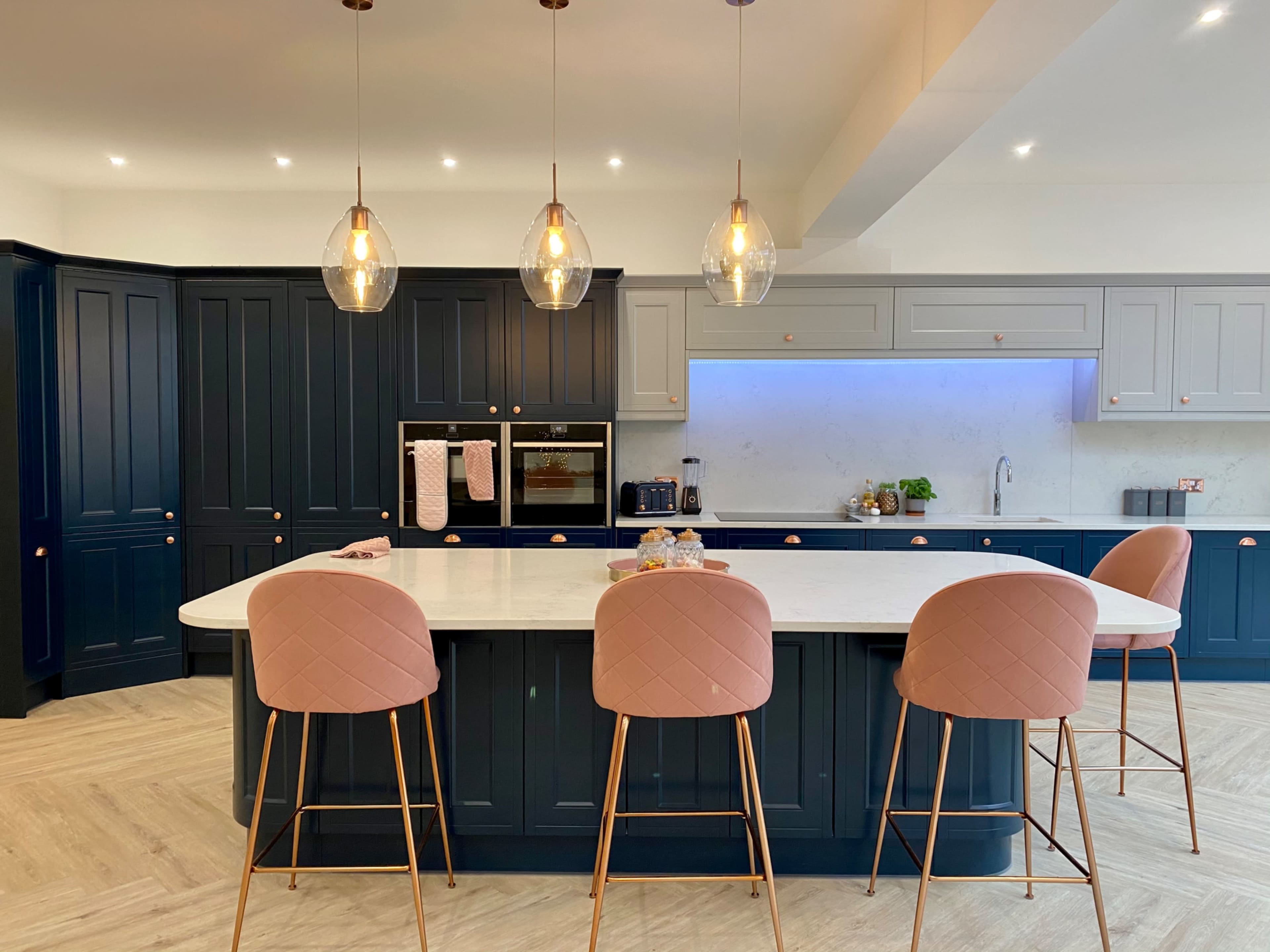 The kitchen features a large island with pink bar stools, dark blue cabinetry, and pendant lights above.
