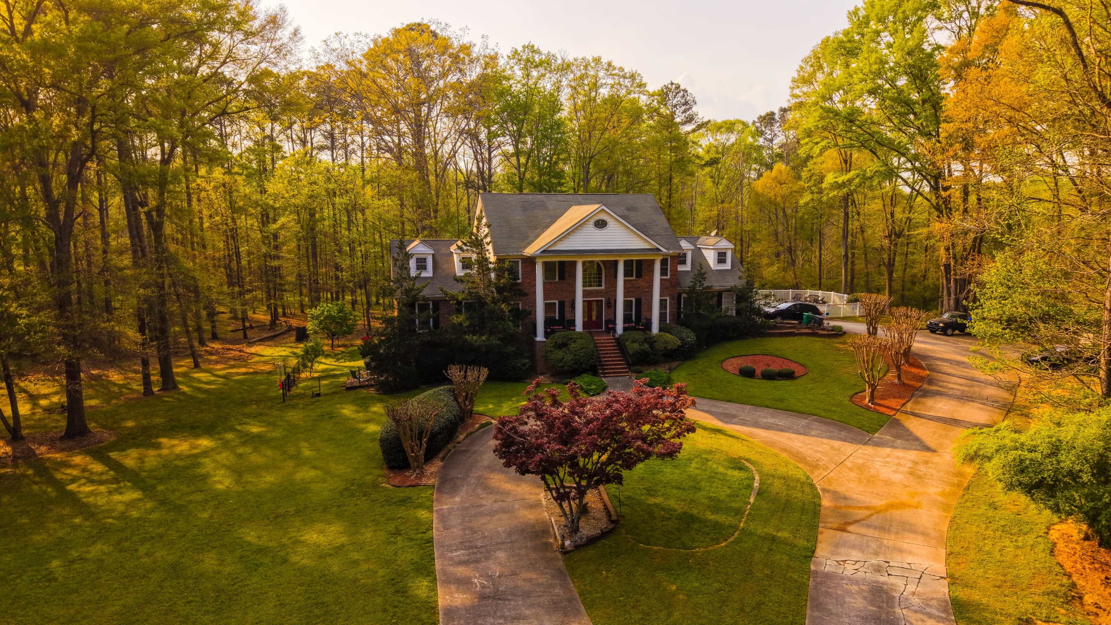 The image shows a large house situated in a forested area, with a circular driveway and landscaped yard surrounded by trees.