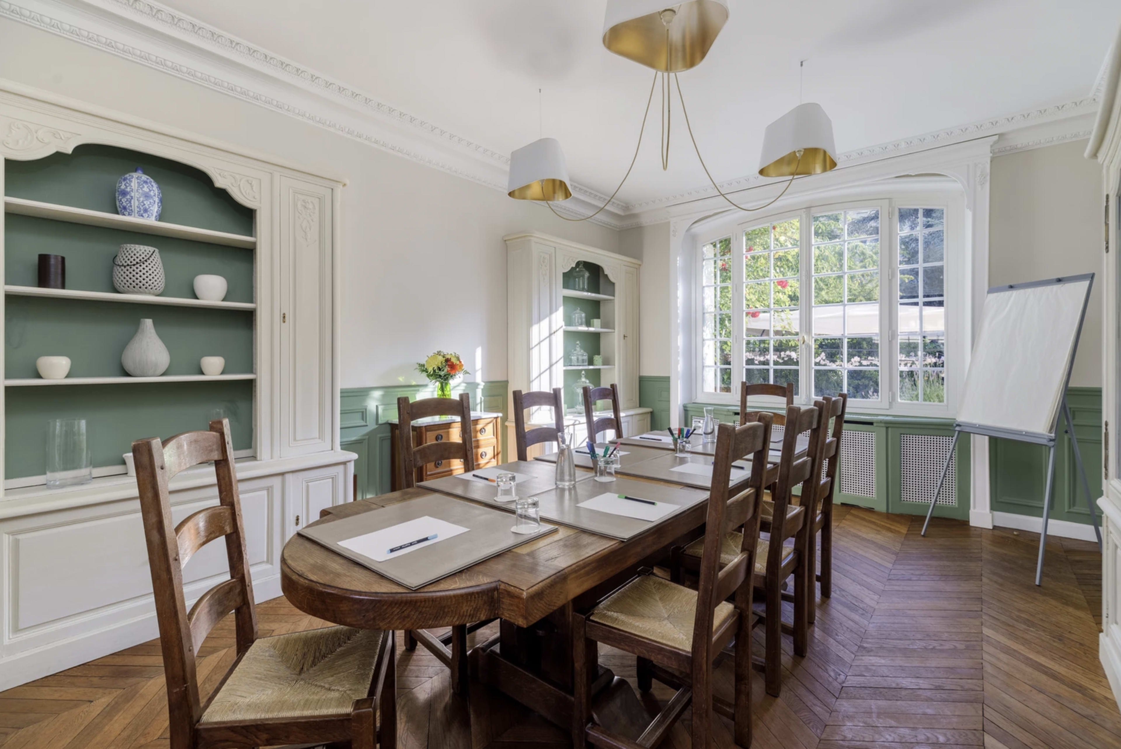 A well-furnished meeting room features a wooden table surrounded by chairs, with a large window allowing natural light and shelves displaying decorative items.