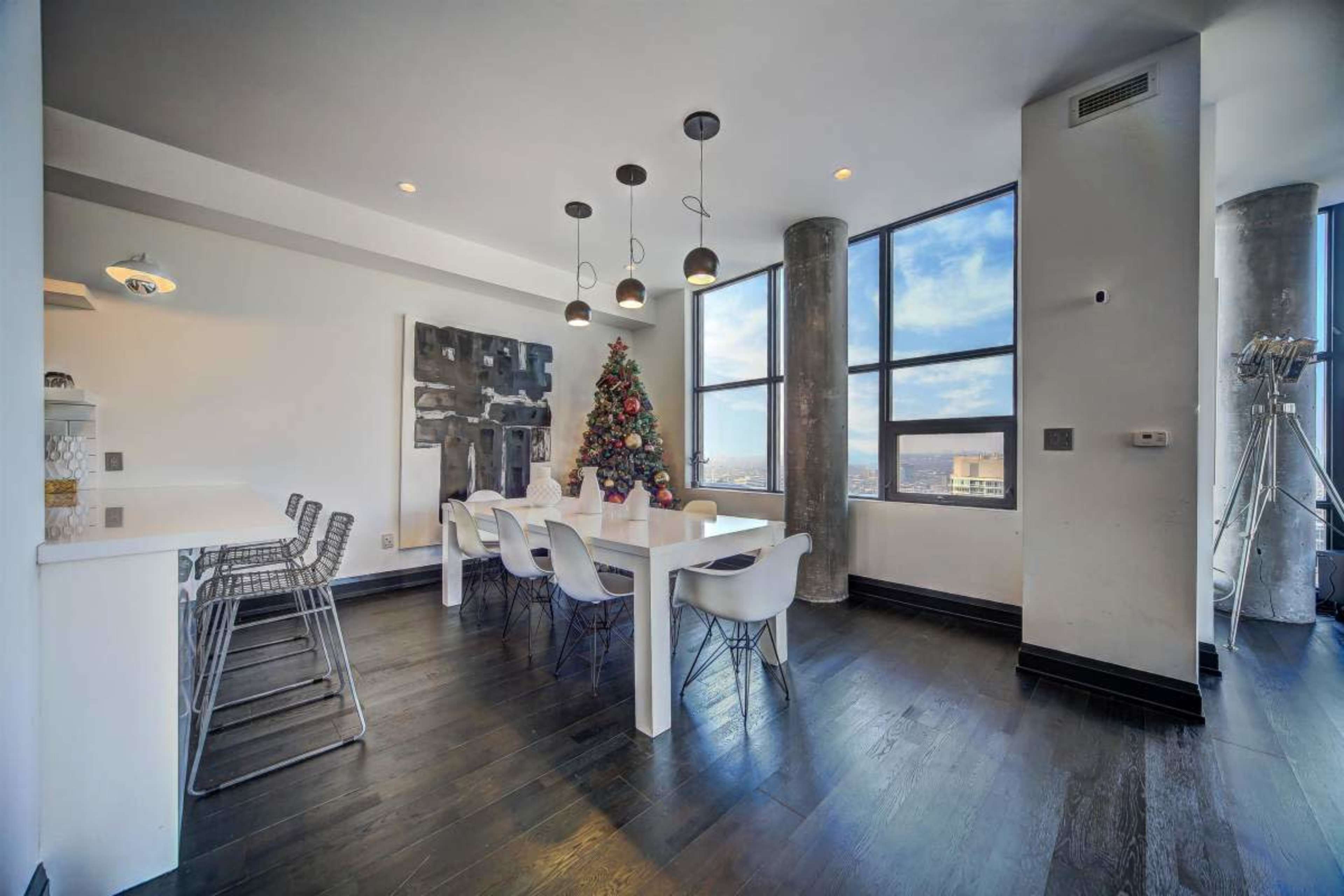 The image shows a modern dining area with a Christmas tree centerpiece, large windows that provide city views, and a combination of white and dark furnishings.