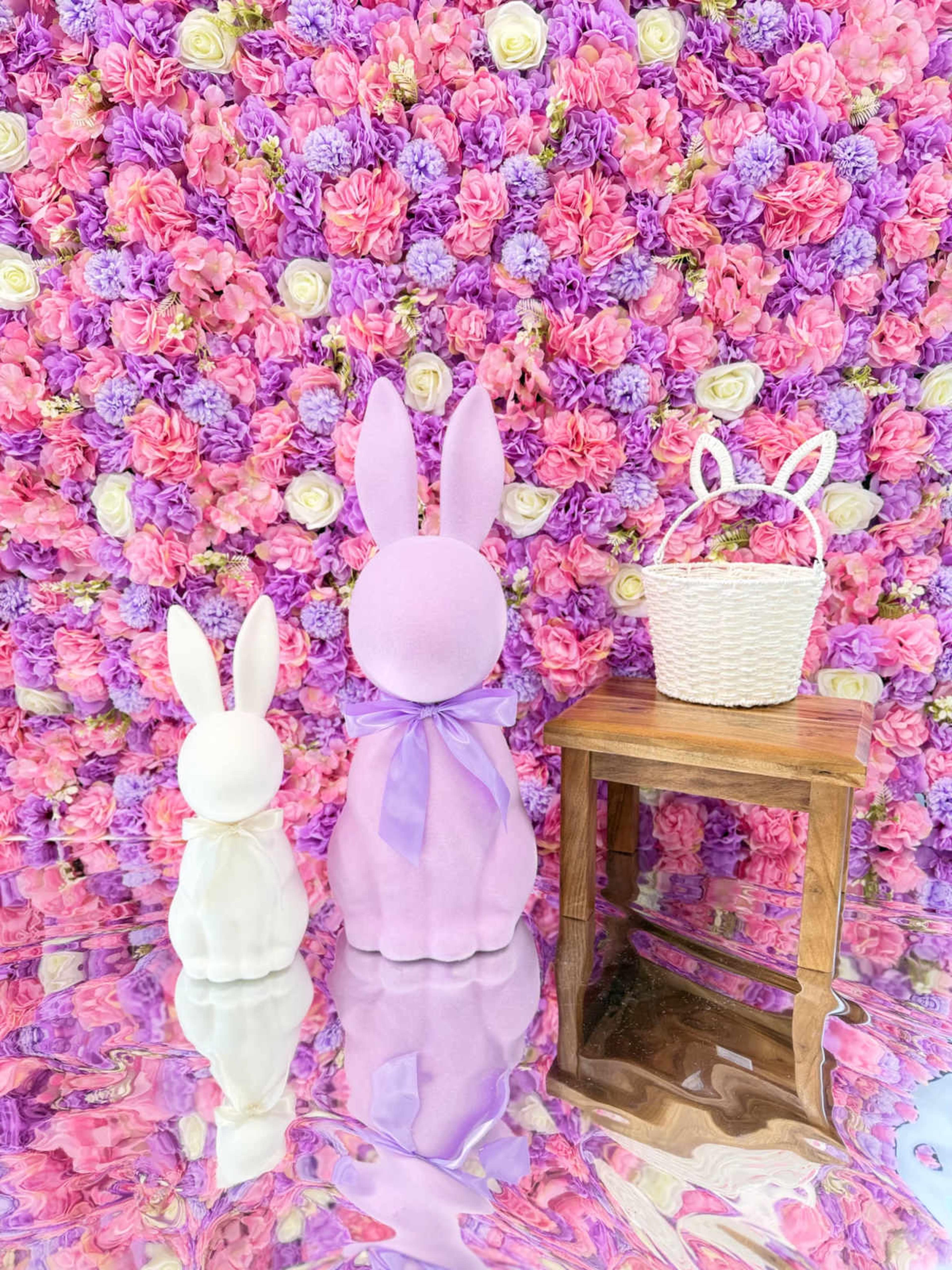 The image features two decorative bunny figurines in front of a colorful floral backdrop, alongside a small wicker basket on a wooden table.