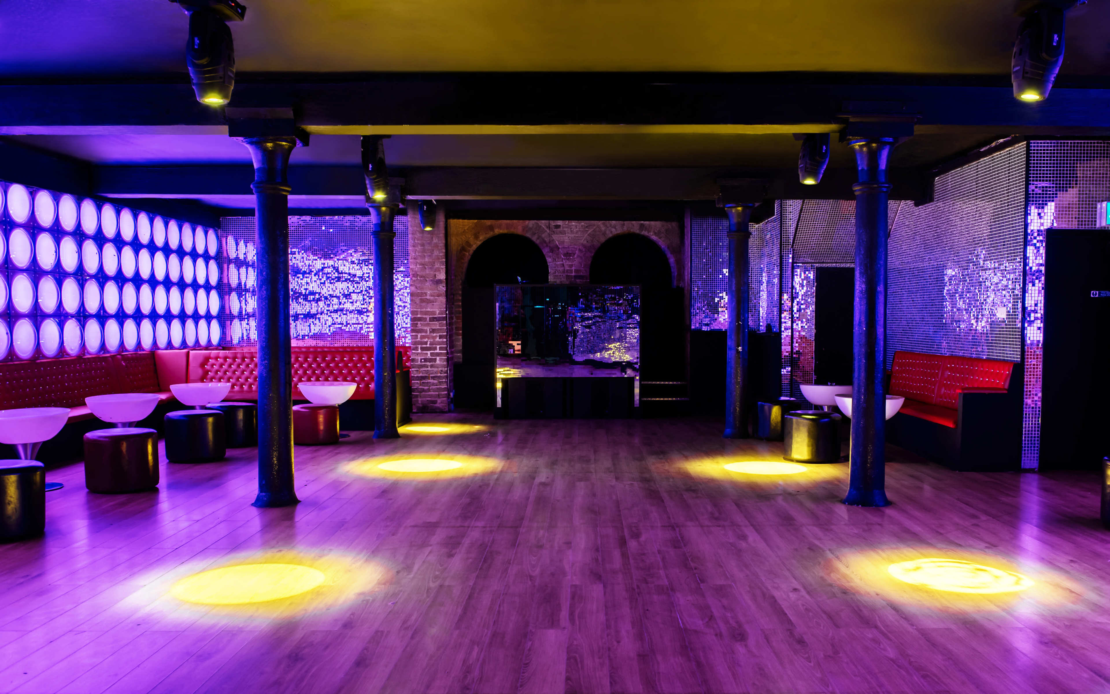 An empty nightclub interior featuring dim lighting, red seating, and a reflective wall.