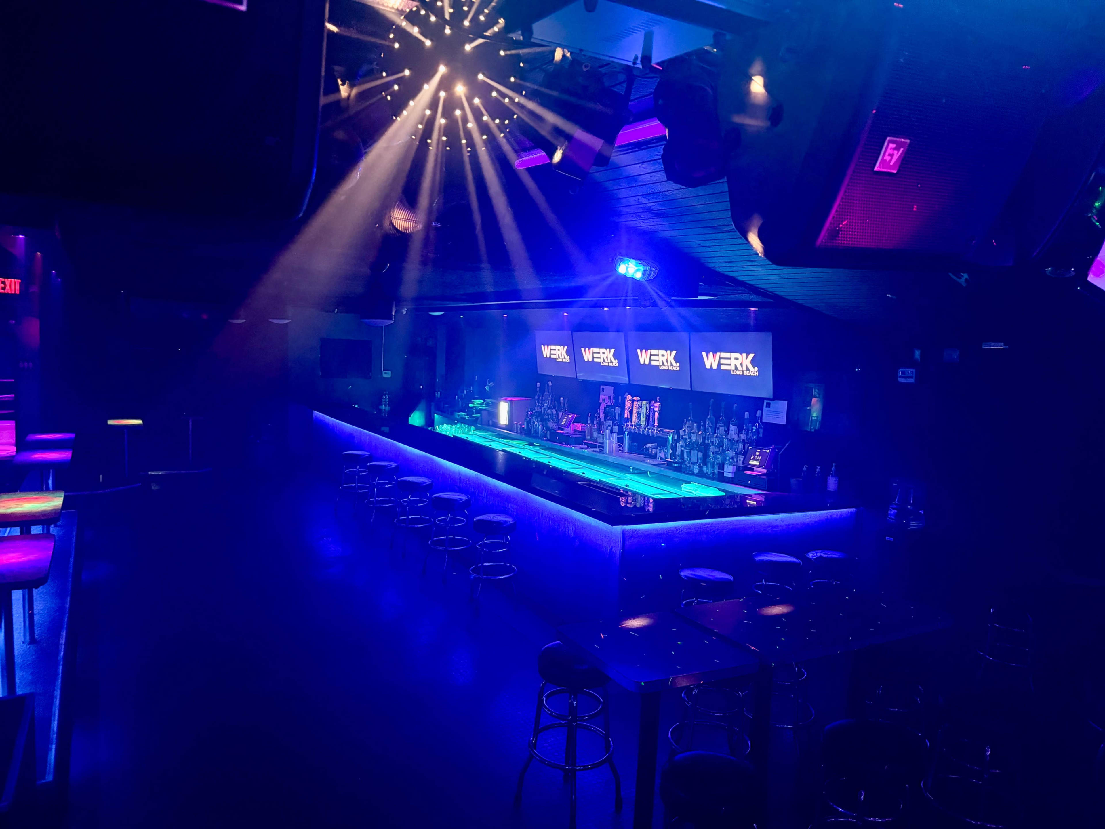The image shows a dimly lit bar with colorful lighting, featuring a central illuminated bar area surrounded by empty tables and chairs.