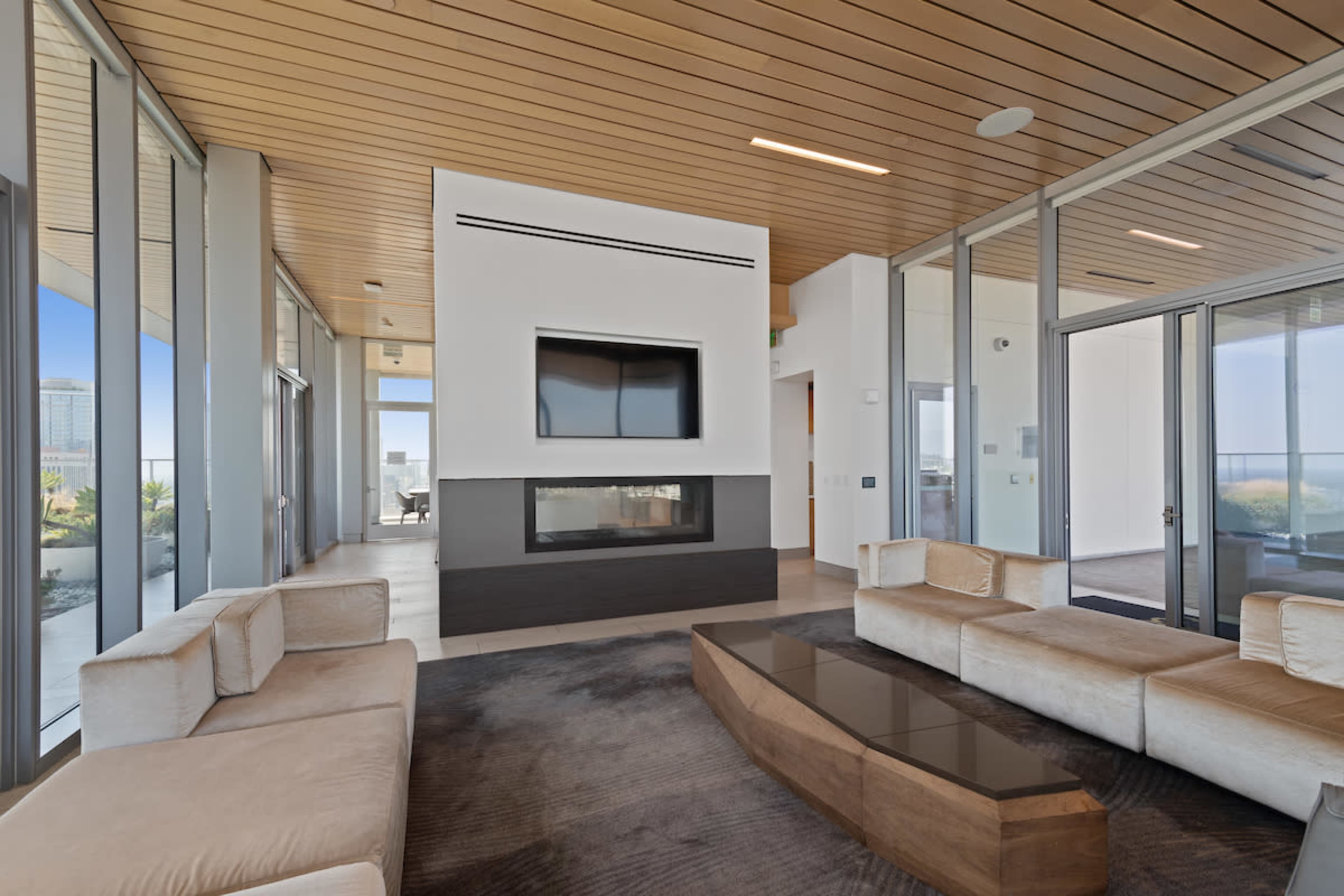 A modern living room with a glass wall, a central coffee table, a fireplace, and a mounted television.