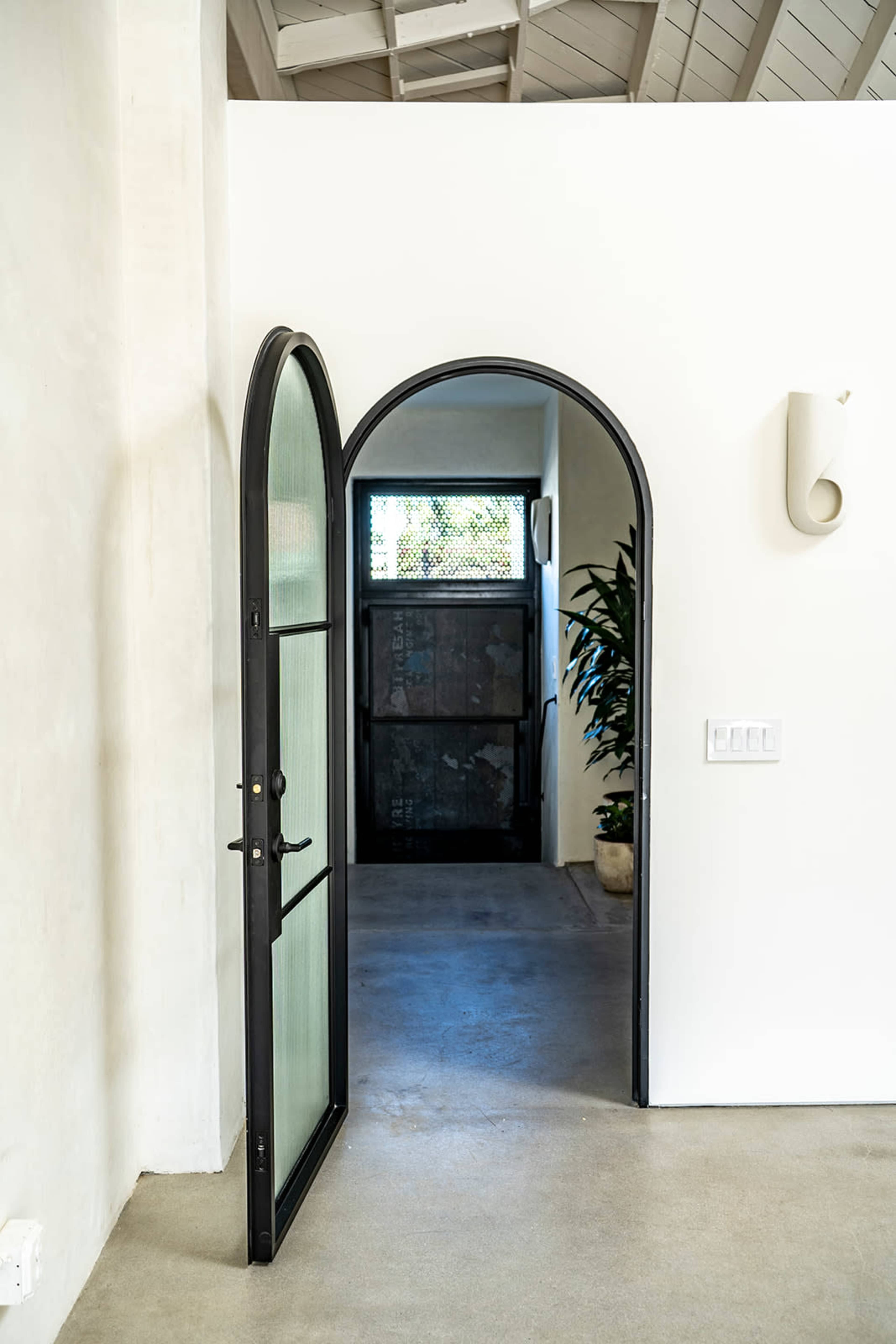 An arched black metal door stands open, revealing a dimly lit indoor space with a staircase and a potted plant beside it.
