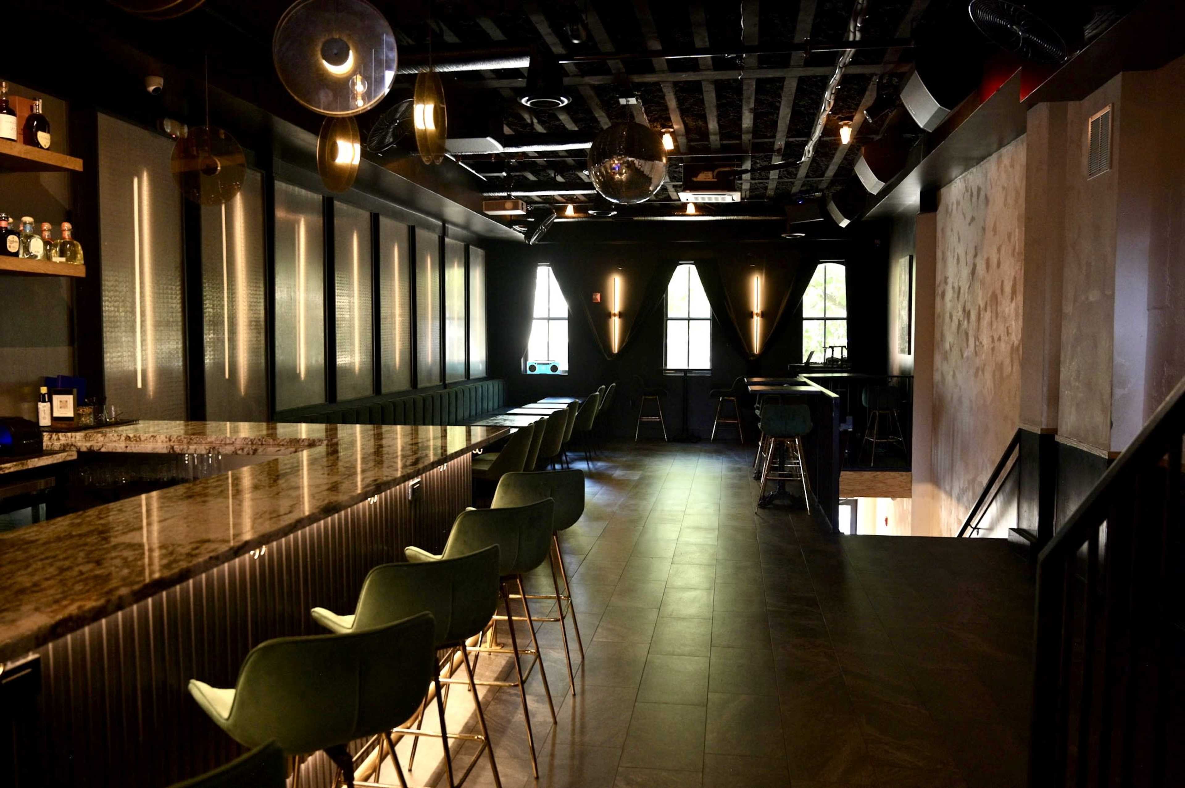 The image shows a dimly lit bar area with a long granite counter, green stools, and modern light fixtures against a dark backdrop.