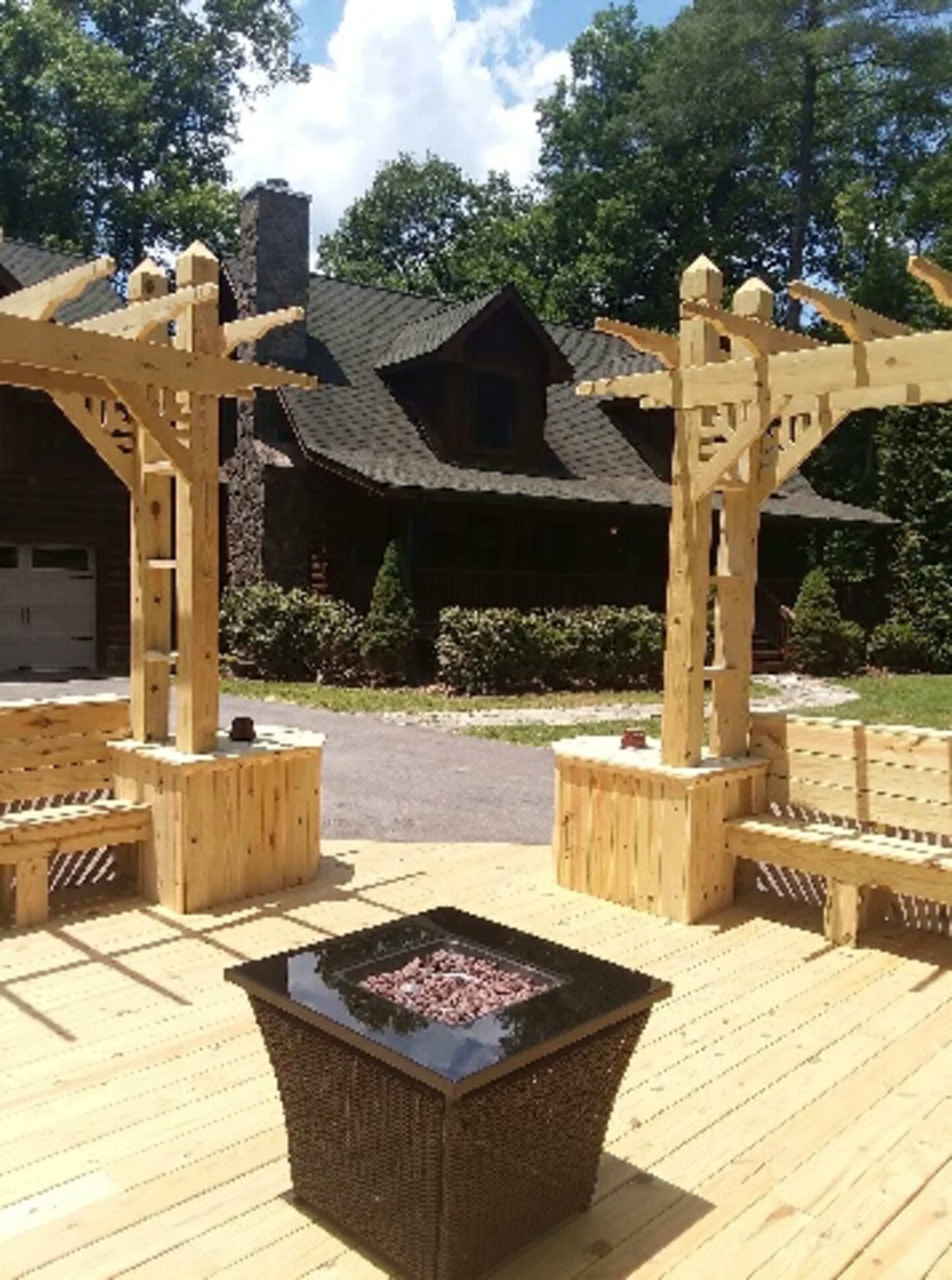 A wooden deck with a fire pit surrounded by benches and two wooden pergolas, set against a house and trees in the background.