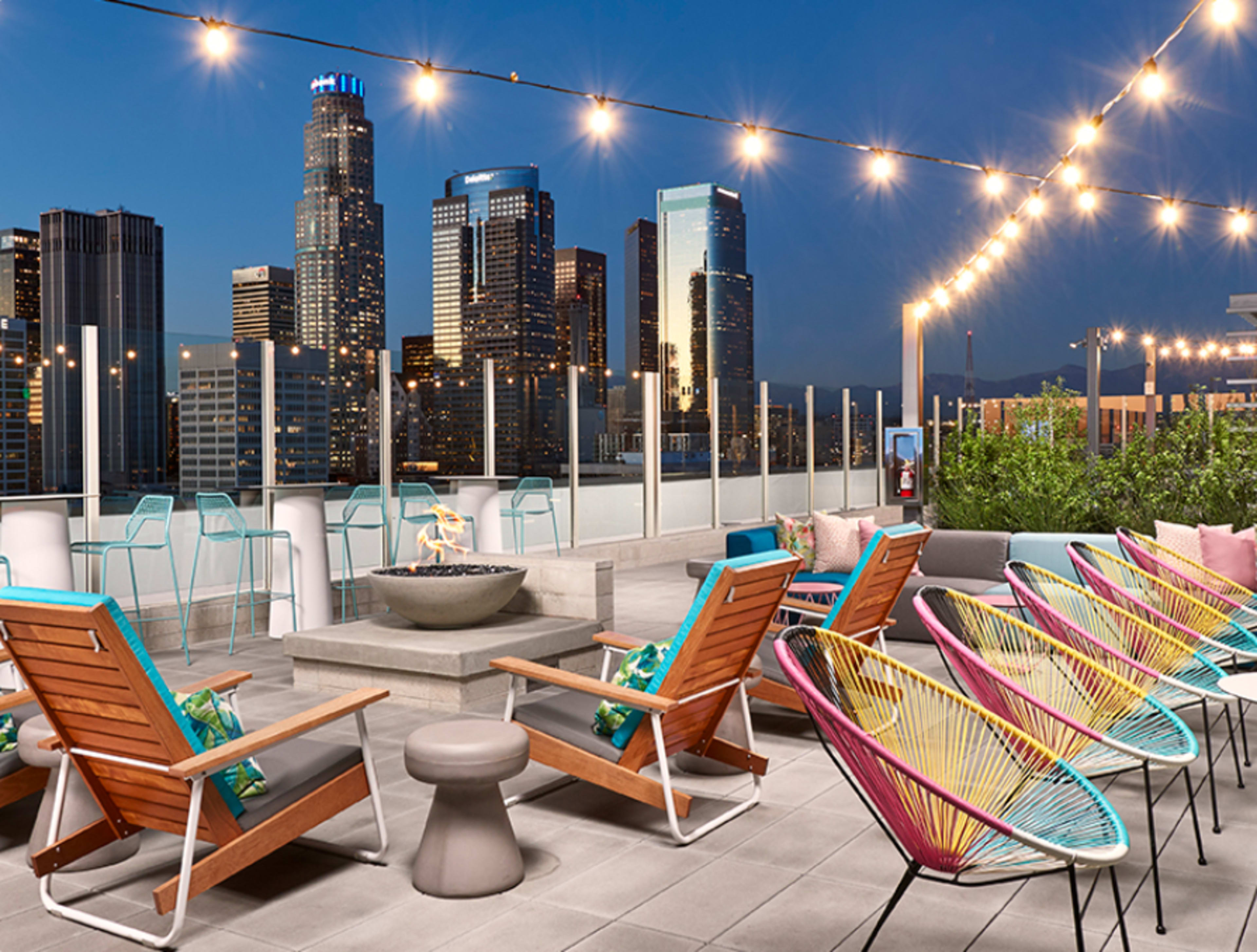A rooftop terrace with colorful seating, a fire pit, and a skyline view of tall buildings illuminated at dusk.