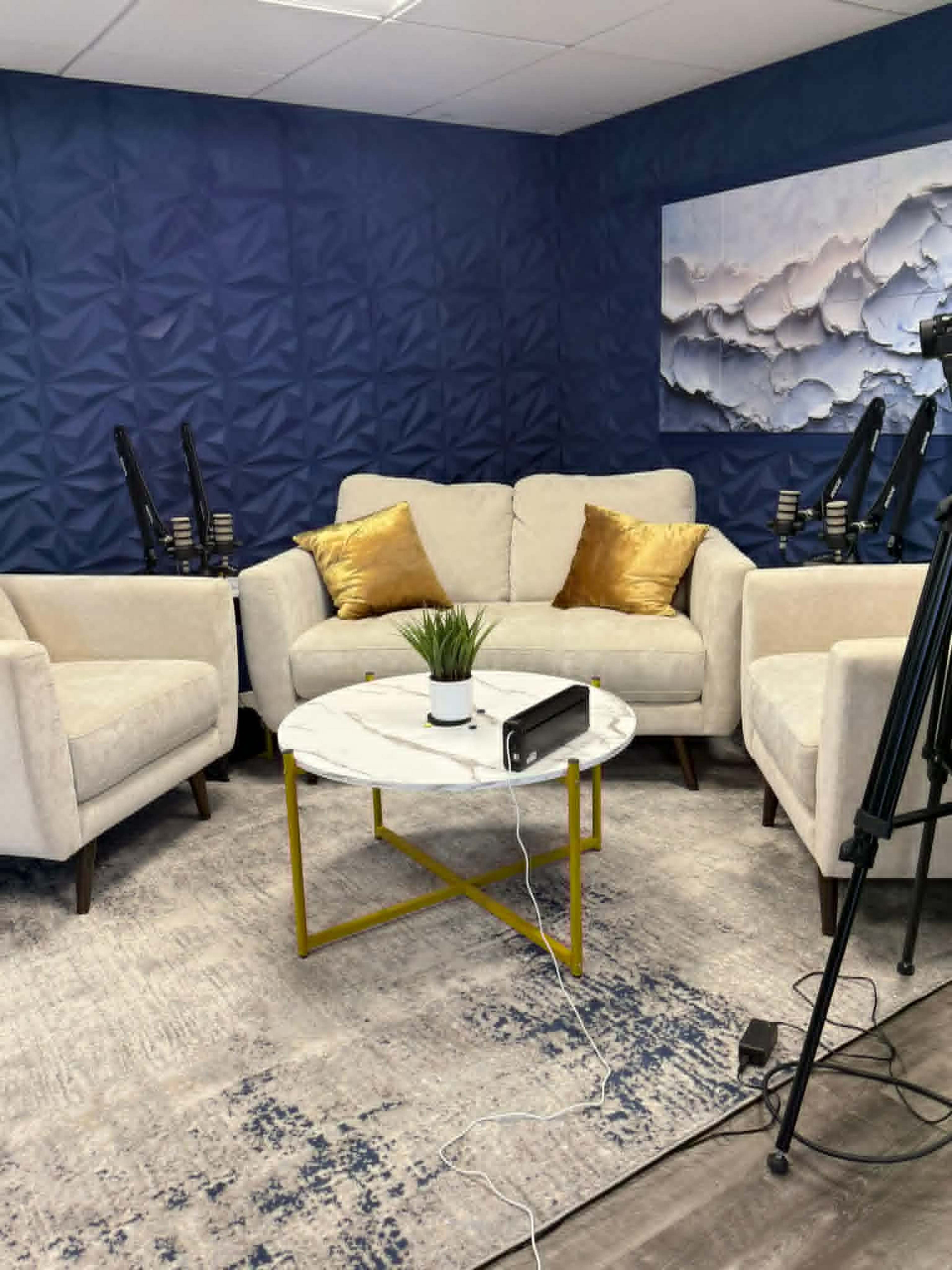 A cozy seating area features two cream-colored chairs and a sofa with gold accent pillows, arranged around a small marble-topped coffee table with a potted plant on top, set against a blue textured wall.