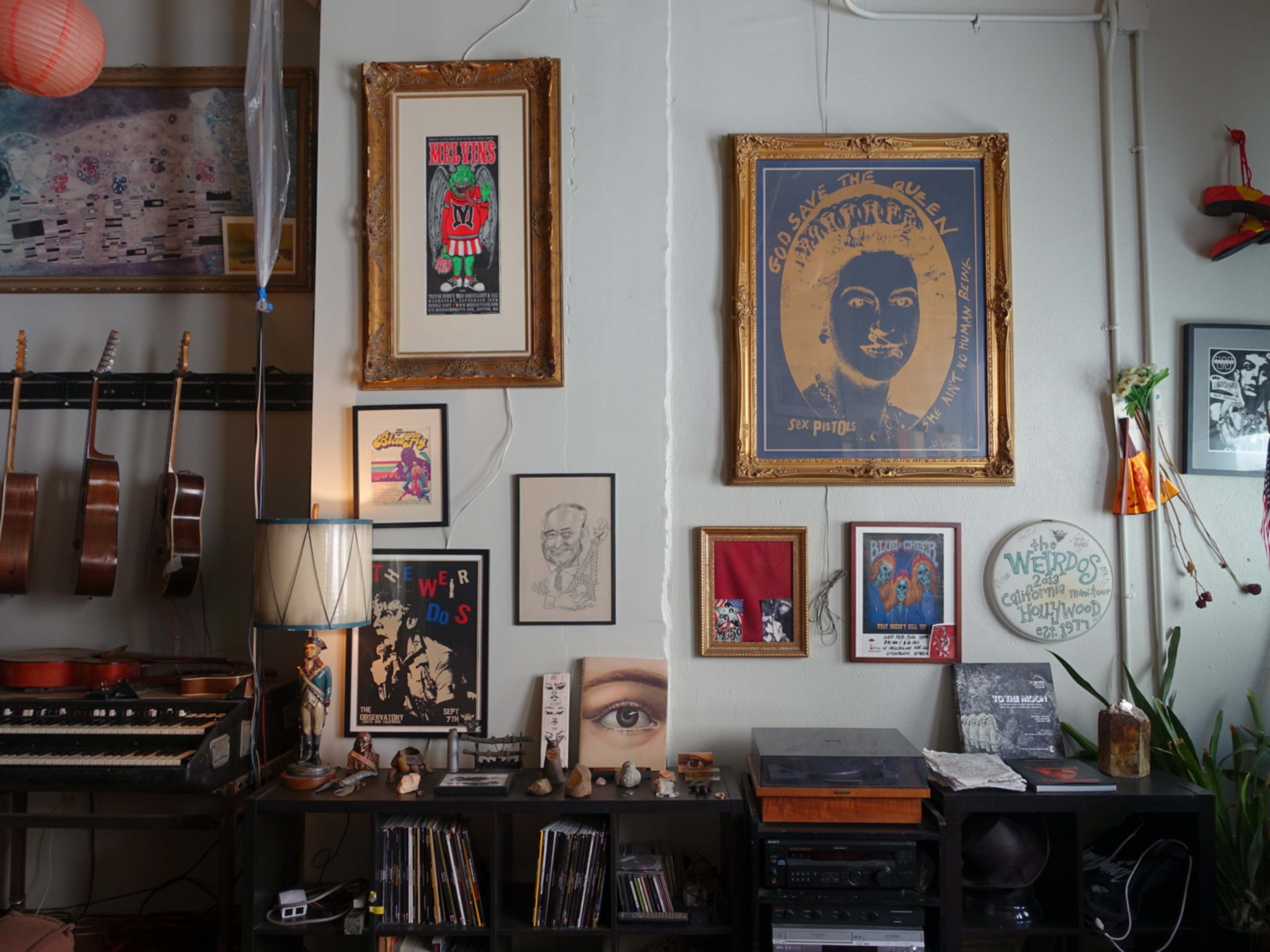 The image shows a wall adorned with various framed artworks, posters, and photographs, accompanied by a collection of musical instruments and a lamp on a shelf below.
