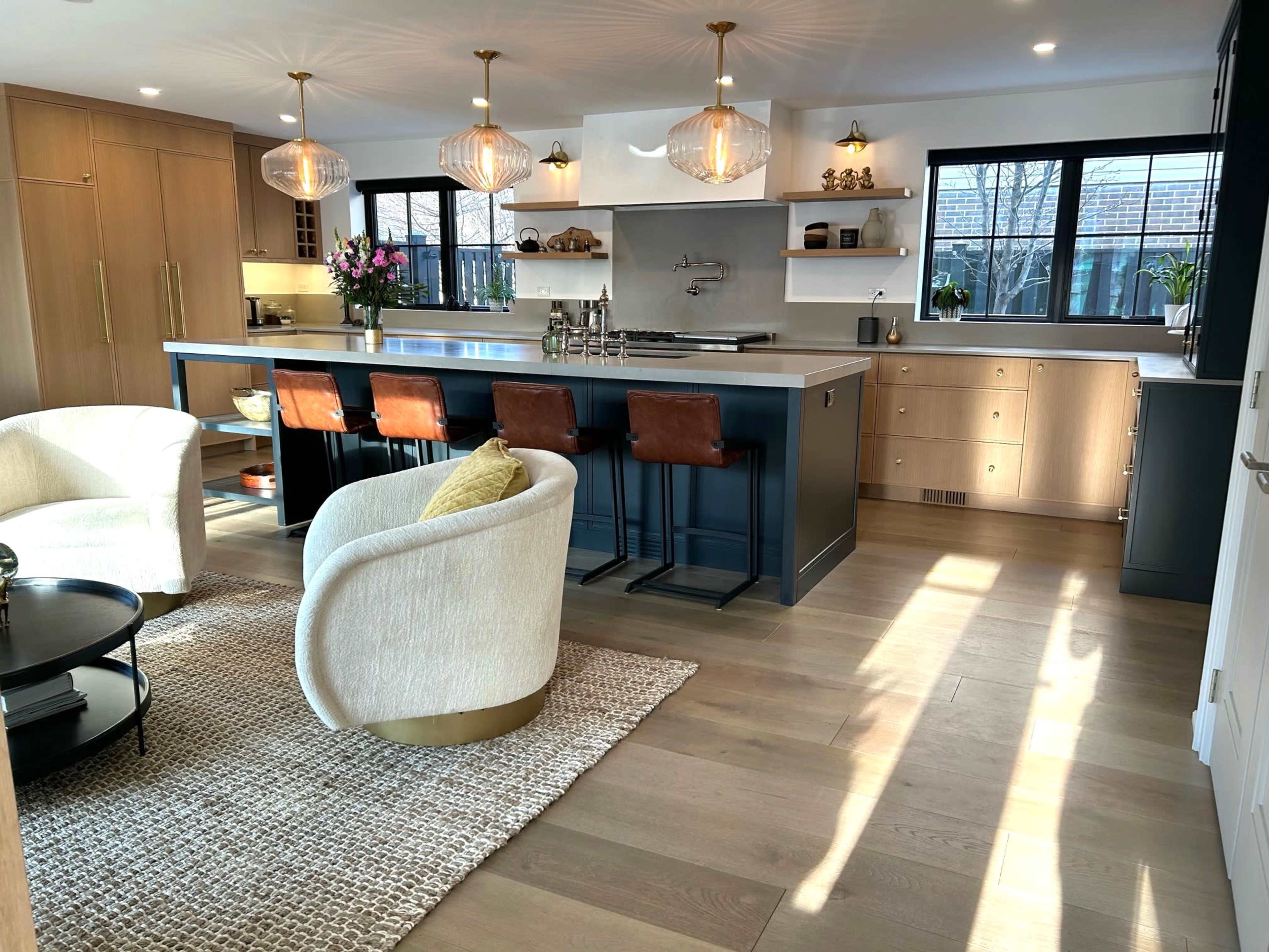 The image shows a modern kitchen and living area with a large island, bar stools, and natural light streaming through the windows.